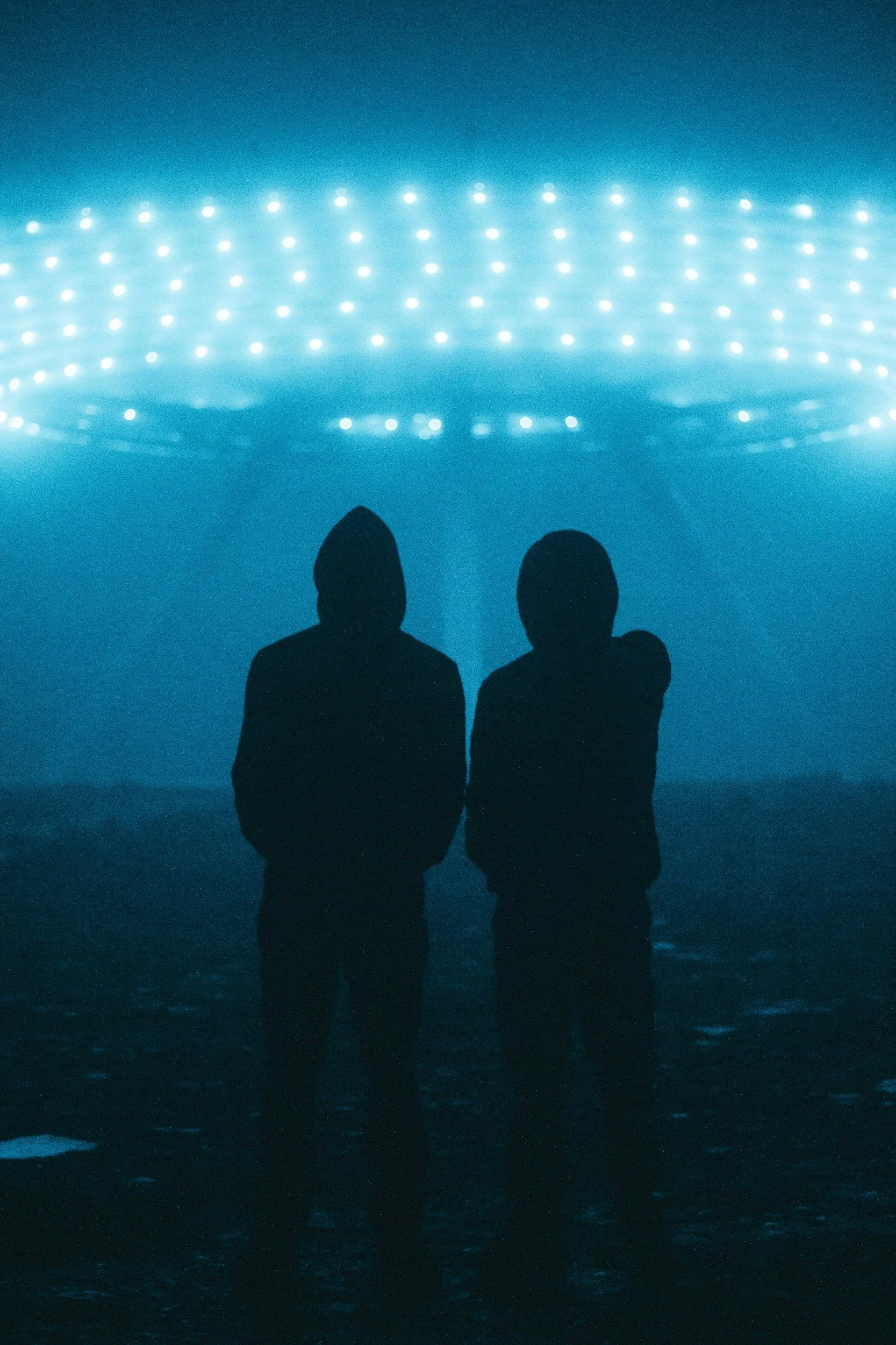 Two people standing in the ocean with a backdrop of bright, colorful light installation in the sky at dusk.