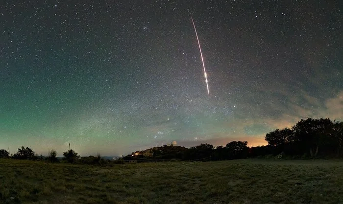 A bright fireball streaks across a blue-green dawn sky above Kitt Peak, with Orion rising over the 2.1-meter Telescope.
