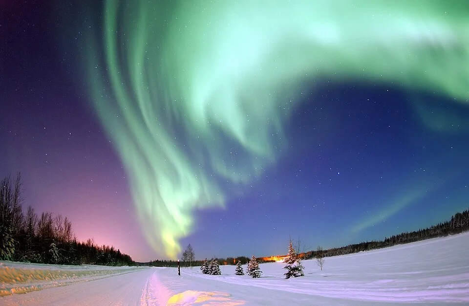 Northern lights over a snowy landscape with trees and a cloudy sky.