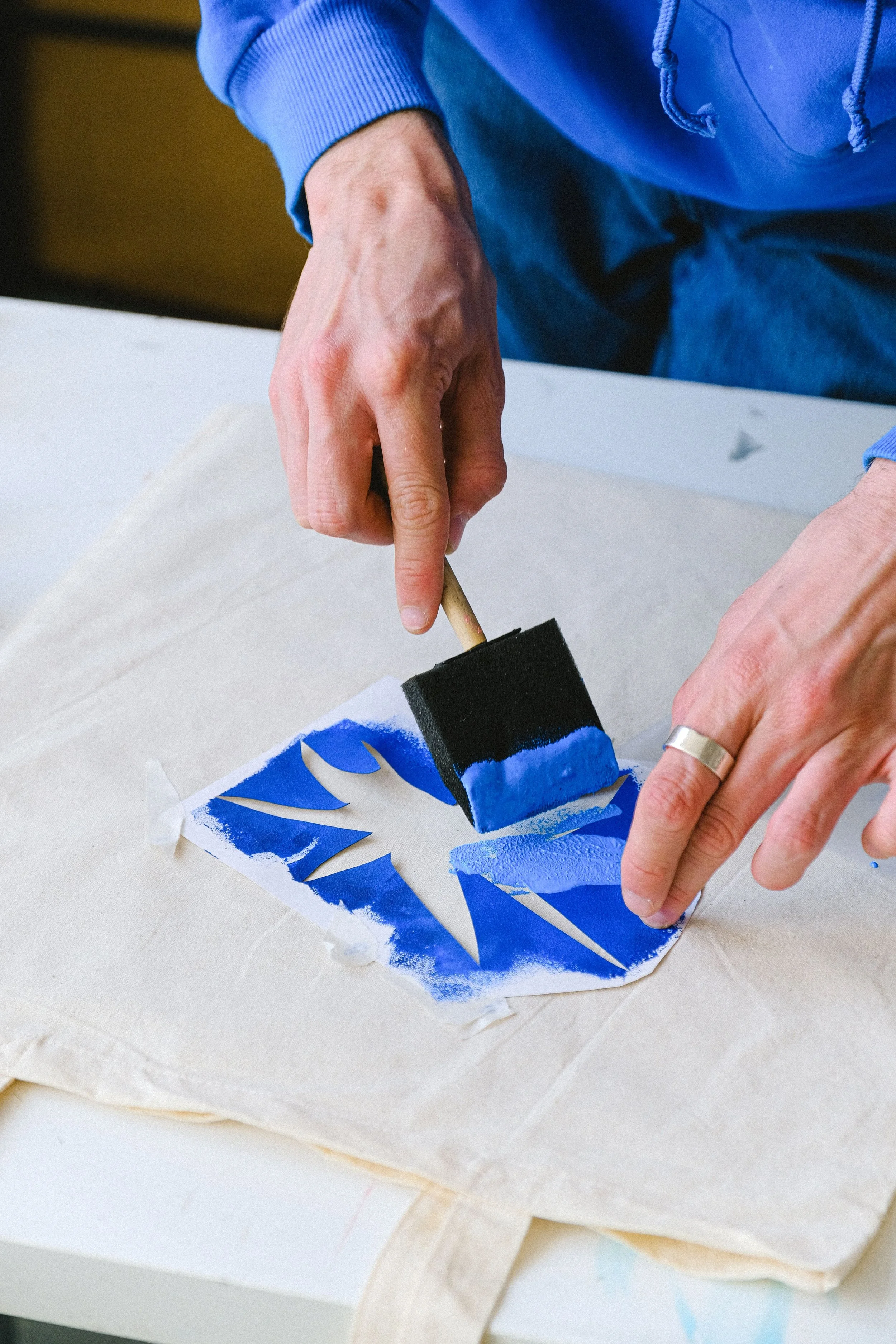 A person using a foam brush to apply blue and black paint to a stencil on paper or fabric.