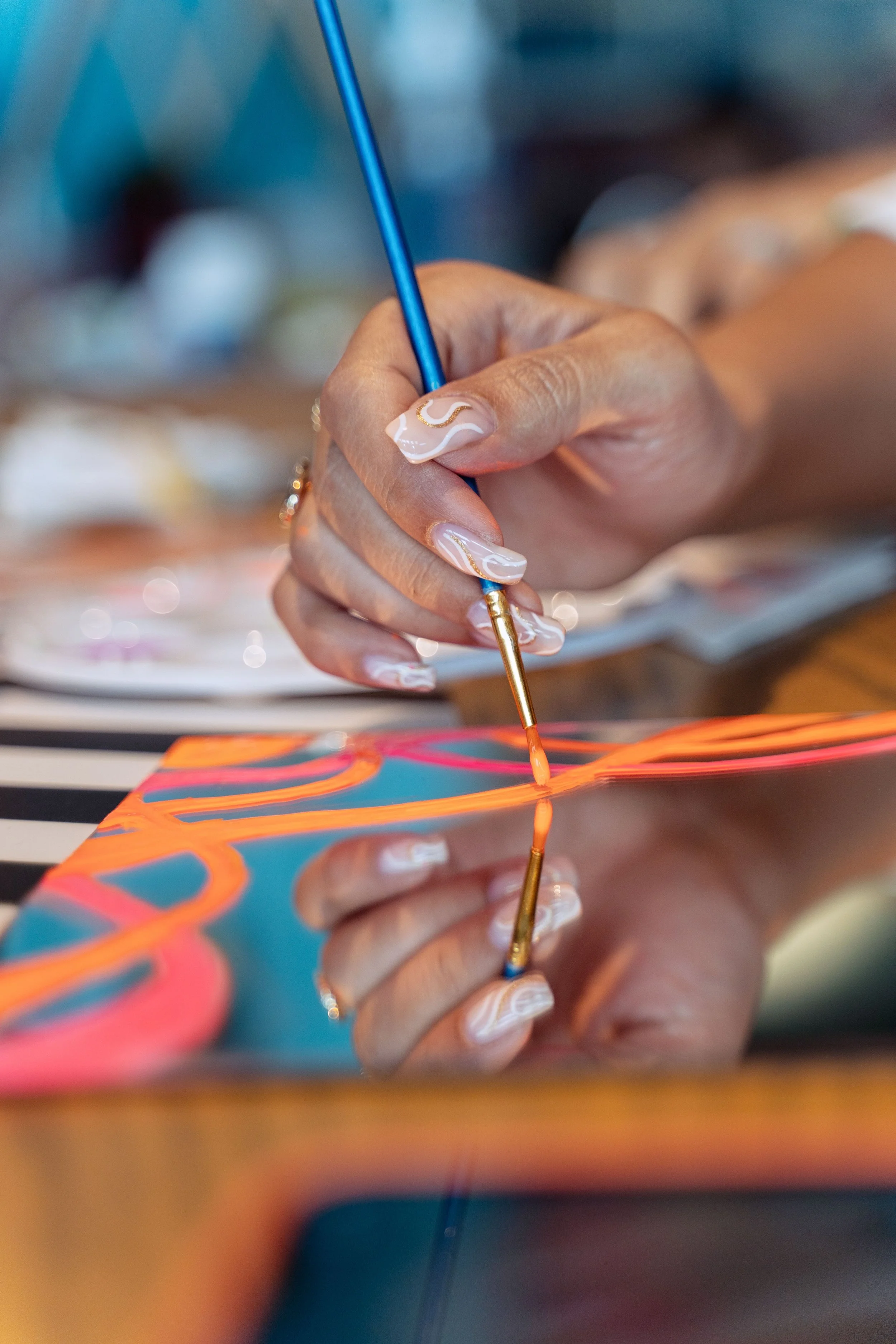 Close-up of a hand with beautifully designed nails holding a paintbrush, painting colorful patterns on a mirrored surface.