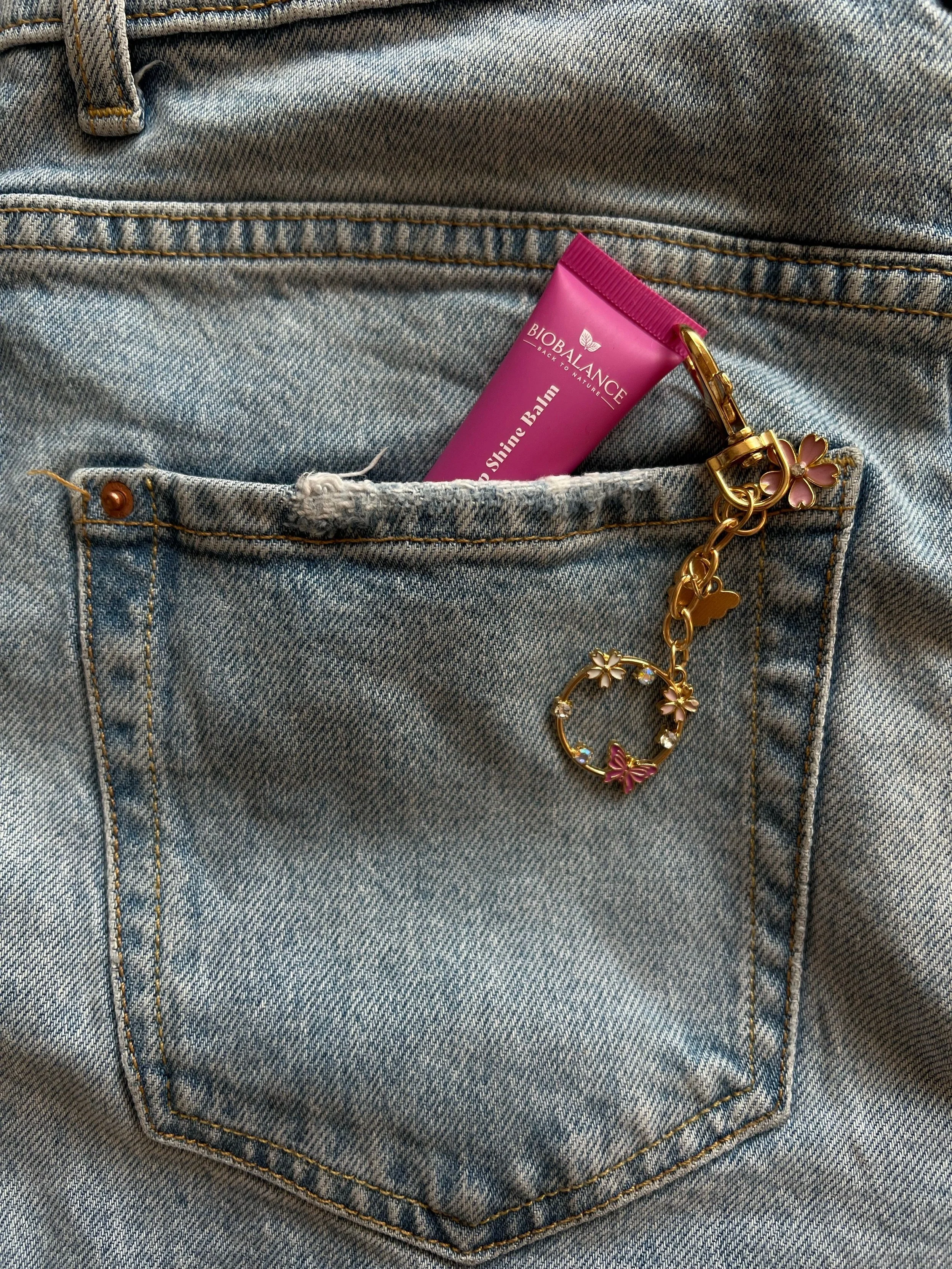 Close-up of a back pocket of blue jeans with a pink tube of Biobalance Shine Balm and a gold chain with pink and white flower charms hanging from the pocket.