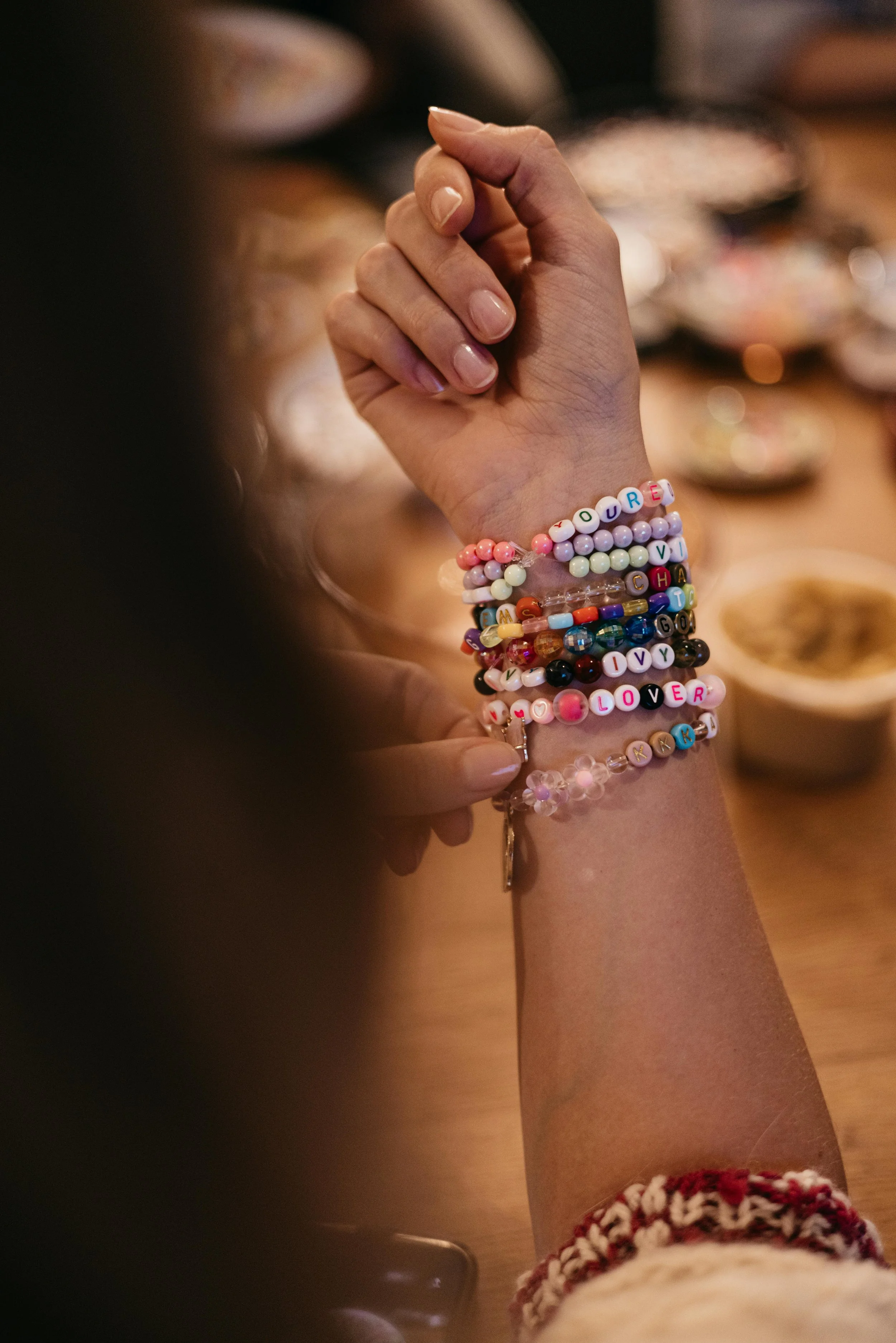 A person's wrist adorned with multiple colorful beaded bracelets, with some beads spelling out words like "LOVE" and "YOU." The person has light skin and their hand is in a partially closed fist. The background is blurred, showing a wooden surface with various items.
