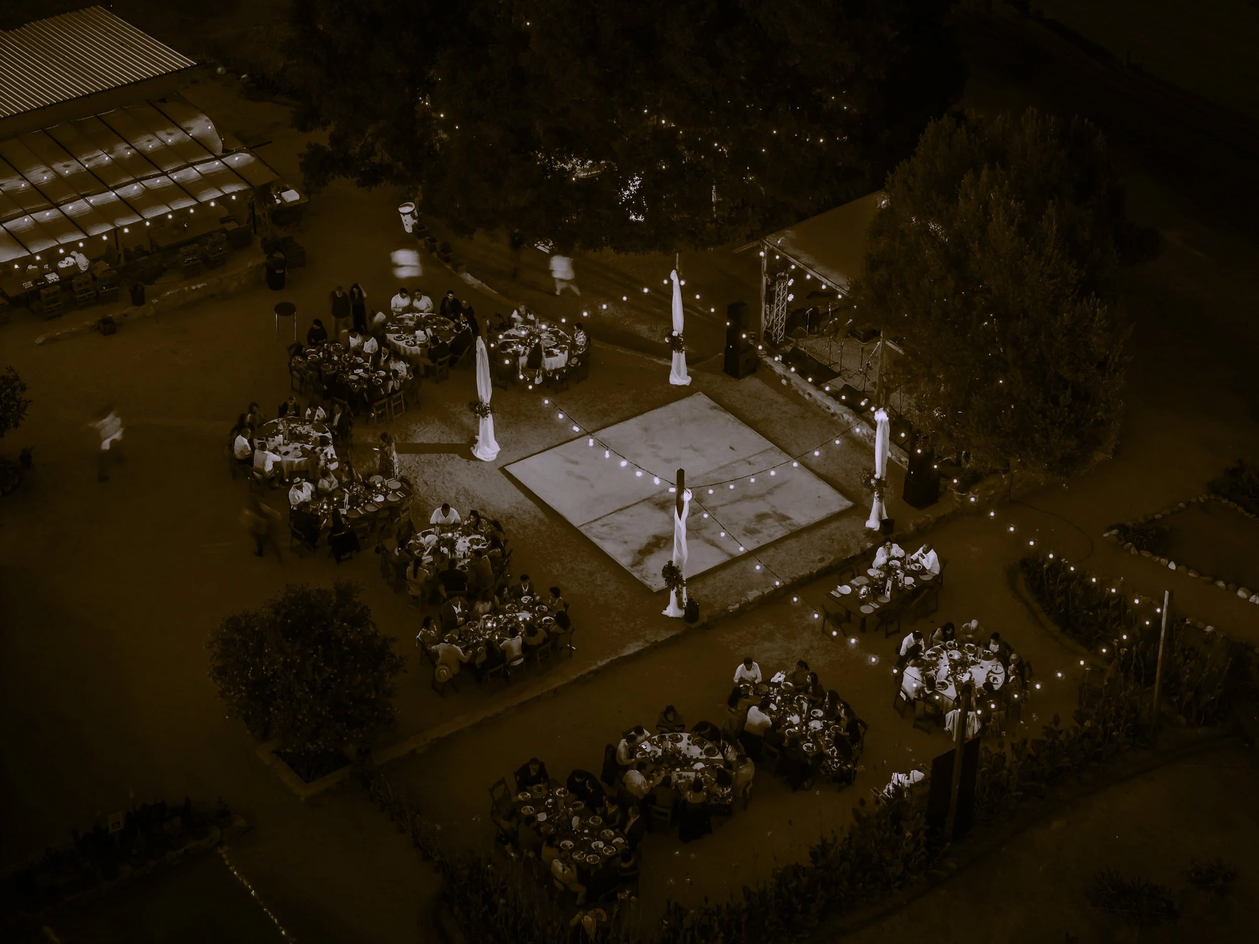Nighttime outdoor social gathering with people sitting at round tables, string lights, a small stage, and an open area with a sand volleyball court.