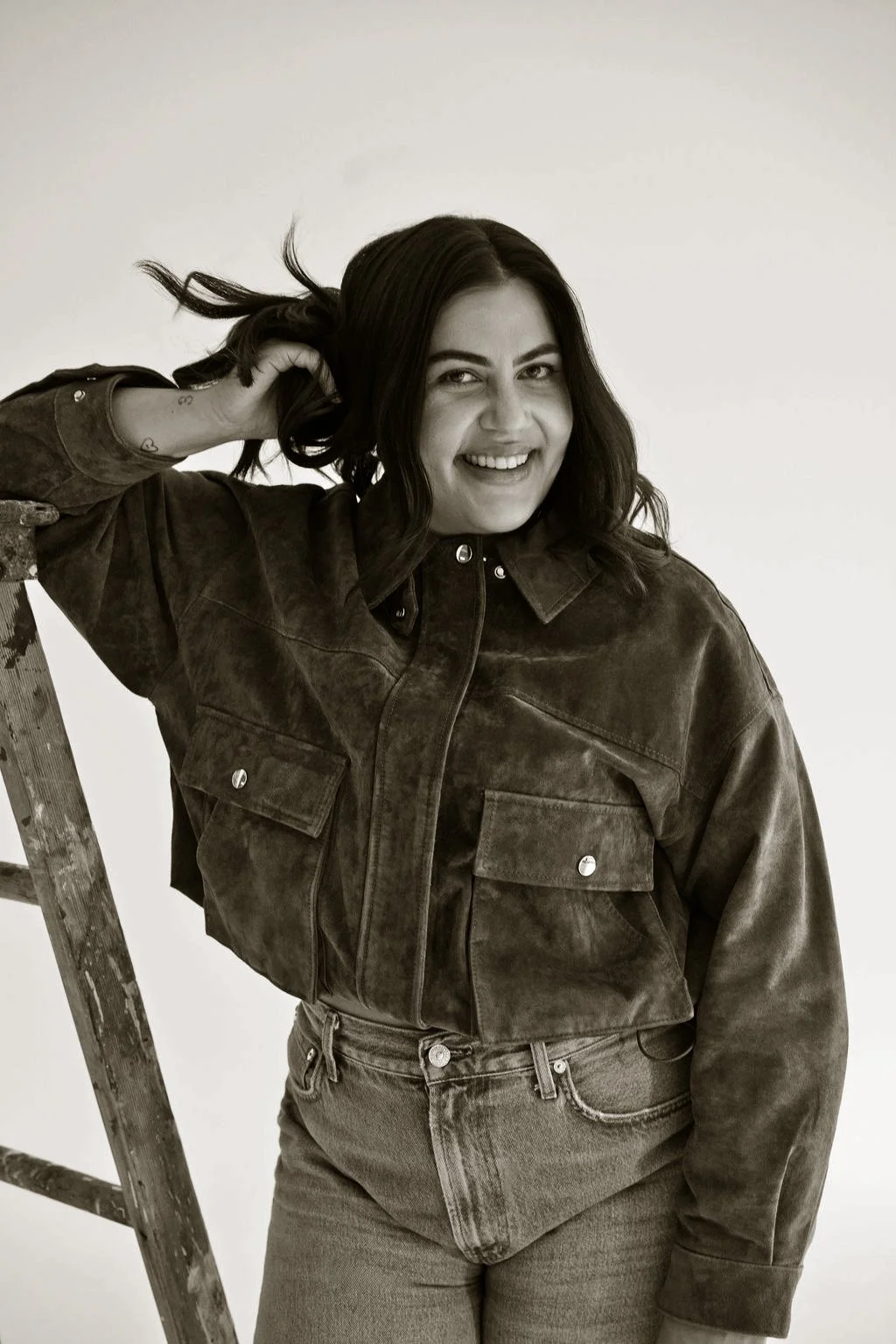A woman with dark hair, smiling and pulling her hair back, standing next to a wooden ladder, wearing a leather jacket and jeans, against a plain light-colored background.