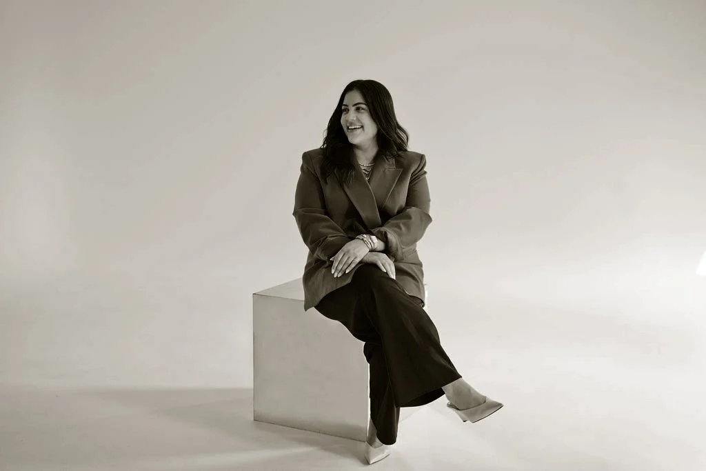 A woman in a blazer and pants sitting on a block, smiling and looking to her left in a photo against a plain background.
