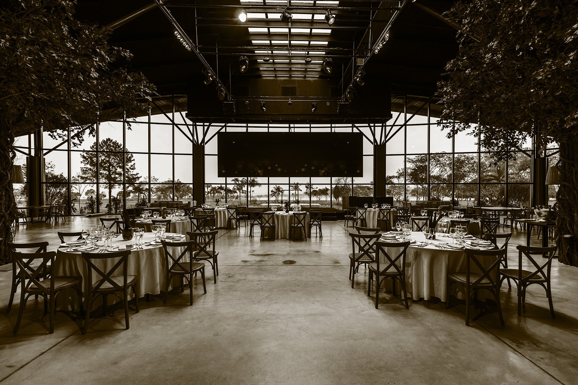 A large, elegant indoor event space with a high ceiling, floor-to-ceiling windows showcasing a view of trees and a water body outside, and a stage area. The room has several round tables with white tablecloths, neatly set with glassware, plates, and utensils. The space is decorated in a modern style with dark accents and natural light streaming in.