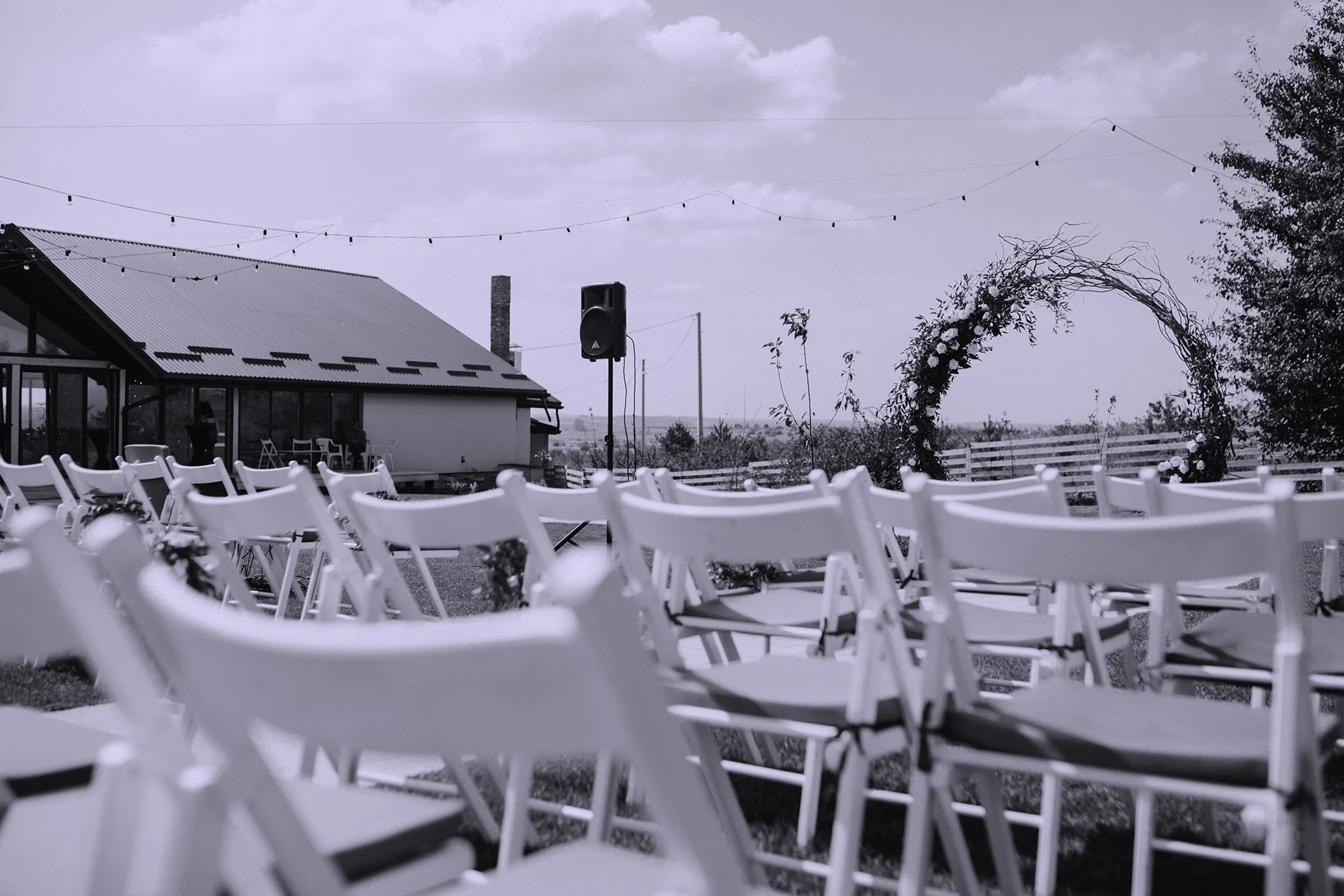 Outdoor event setup with rows of white folding chairs, a building with a metal roof, string lights overhead, and a decorative arch with flowers, under a cloudy sky.