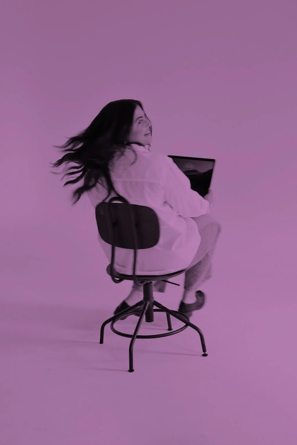 A woman with long hair sitting on a black chair, holding a laptop, with a purple background.