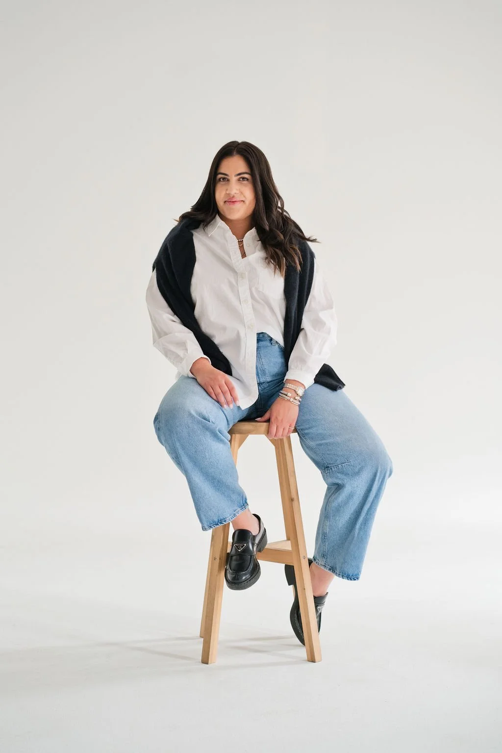 A woman with dark hair sitting on a wooden stool against a white background, wearing a white shirt, light blue jeans, black loafers, and has a black sweater draped over her shoulders.