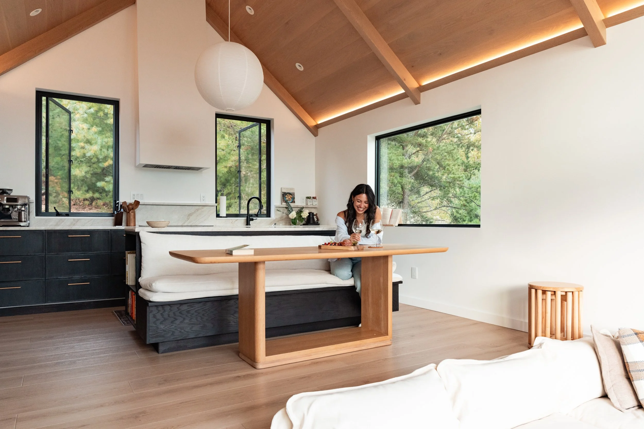 A woman smiling and sitting at a kitchen island with wine glasses and food, in a modern kitchen with large windows and wooden ceiling beams.