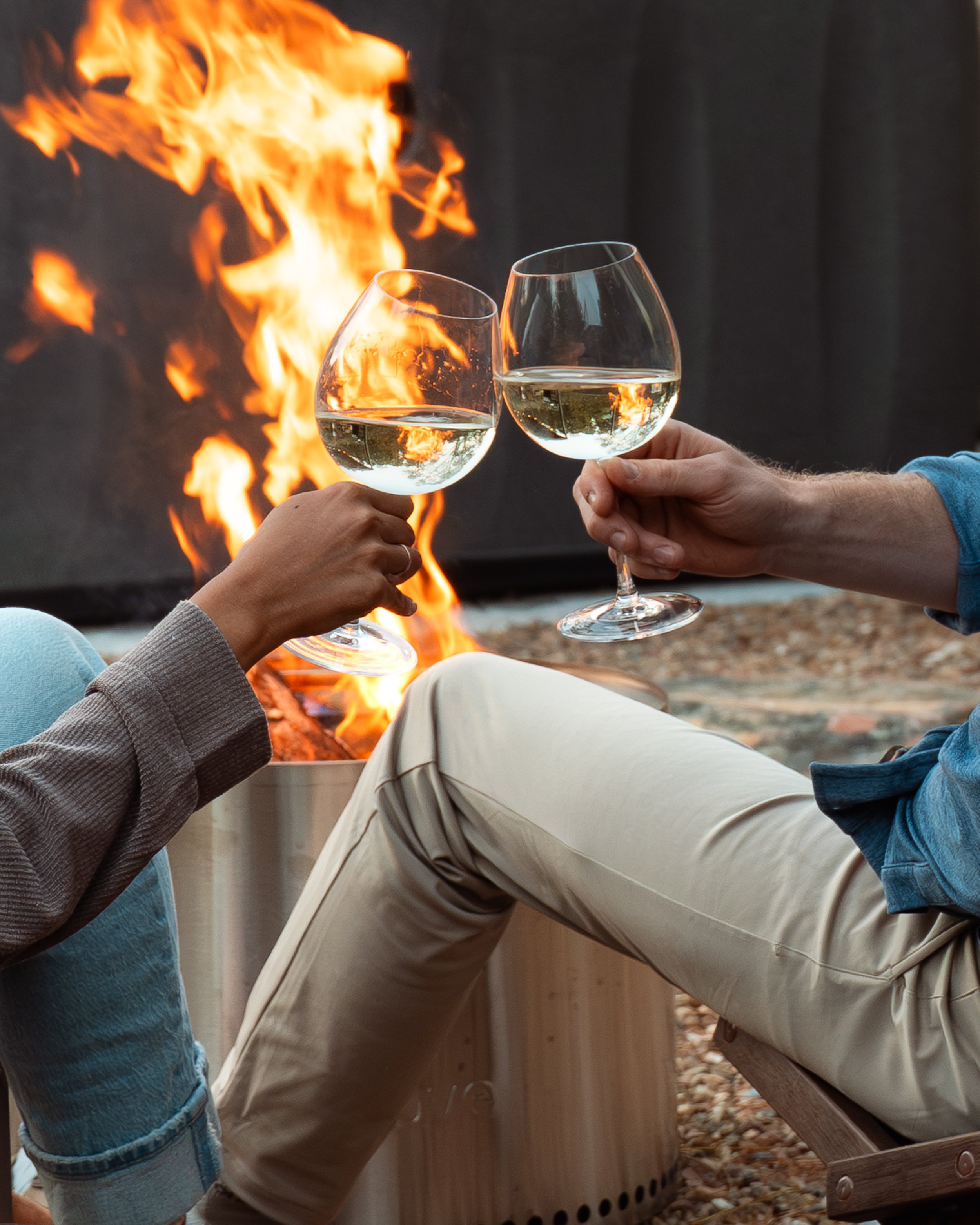 Two people clinking glasses of white wine in front of a fireplace with flames.