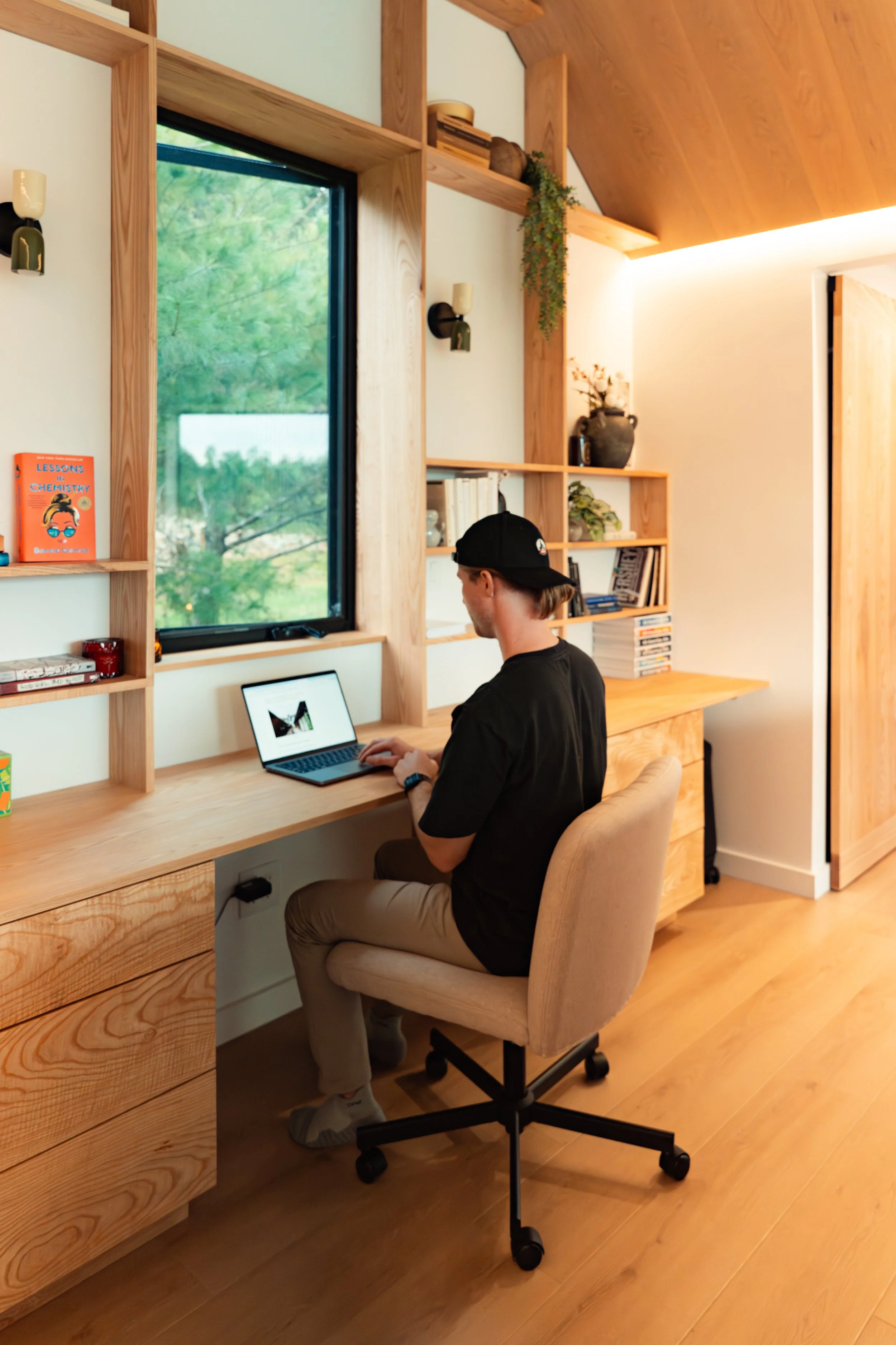 A man sitting at a wooden desk working on a laptop in a room with a large window showing green trees outside. The room has wooden shelves, some decorated with books and plants, and beige walls and flooring.