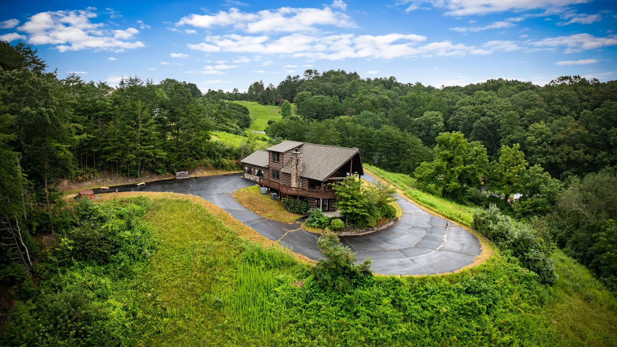 A house situated in a lush green, forested area with a paved driveway and a circular turnaround. The house has a stone chimney and a wooden deck, with some storage or containers around it. The scene is under a partly cloudy sky.