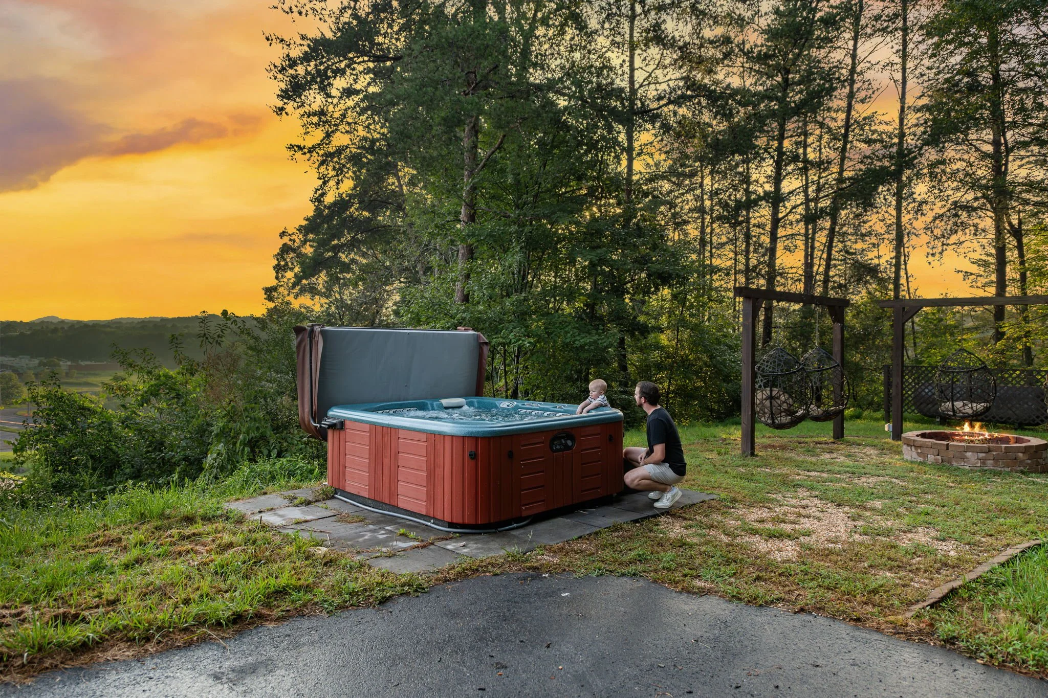 A backyard scene during sunset with a man and a young child near an outdoor hot tub on a paved surface, trees and a sunset sky in the background.