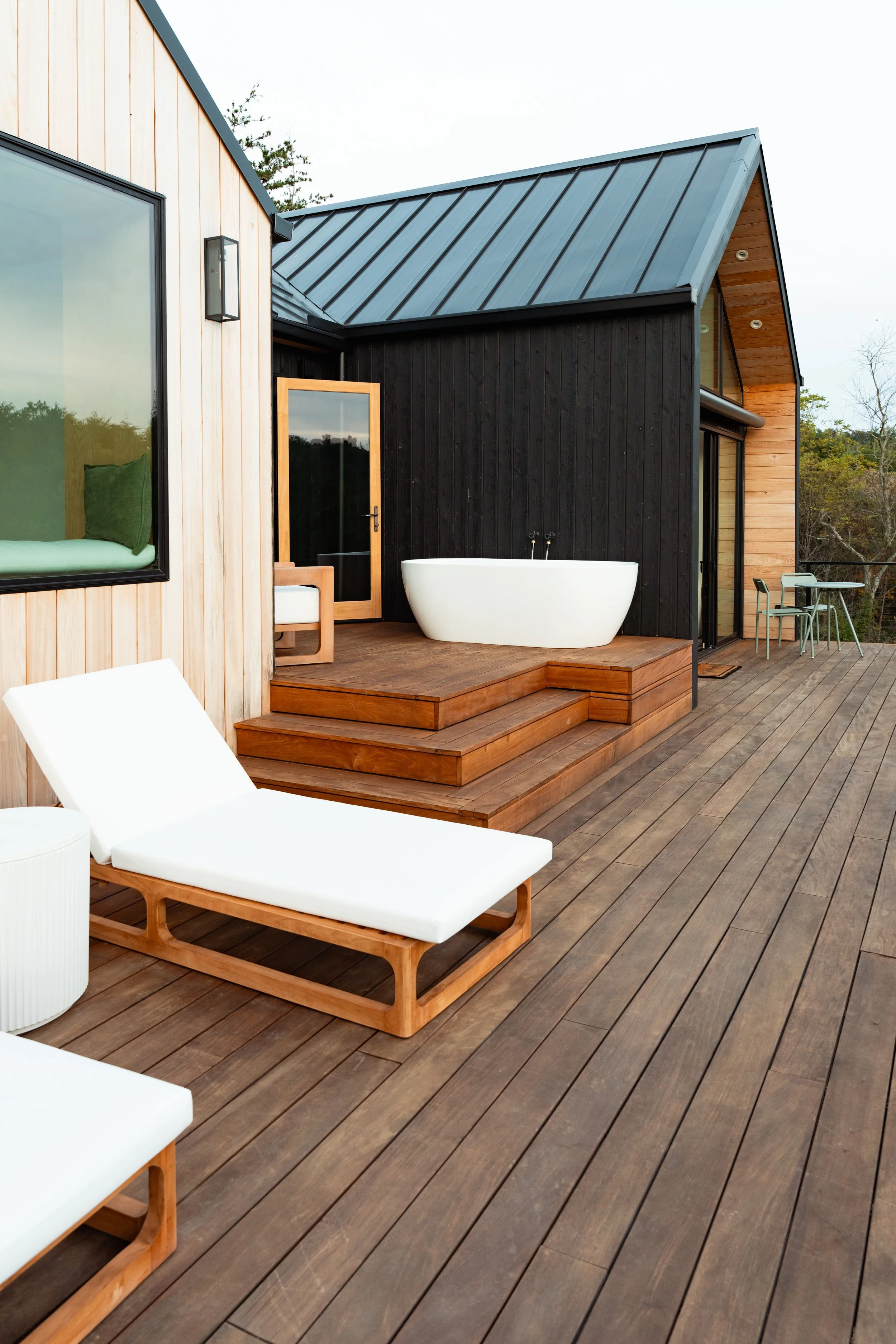Exterior view of a modern house with wooden and black siding, featuring a deck with outdoor furniture and a white bathtub on the deck.