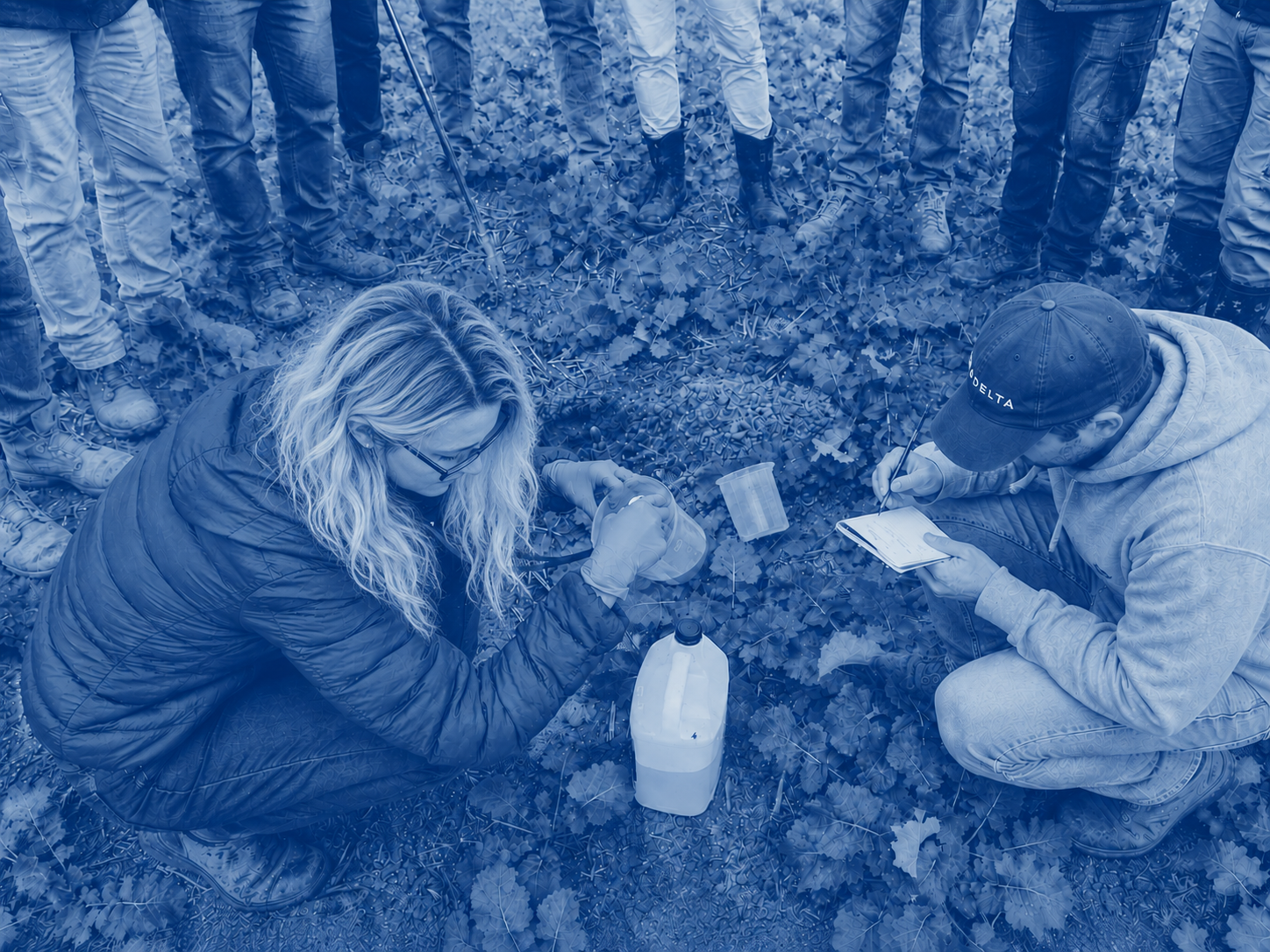 Two researchers collecting plant and soil samples from an agricultural cropping field of leafy plants, one person is kneeling with a plastic container while the other is writing notes on a clipboard; several farmers stand around them observing.