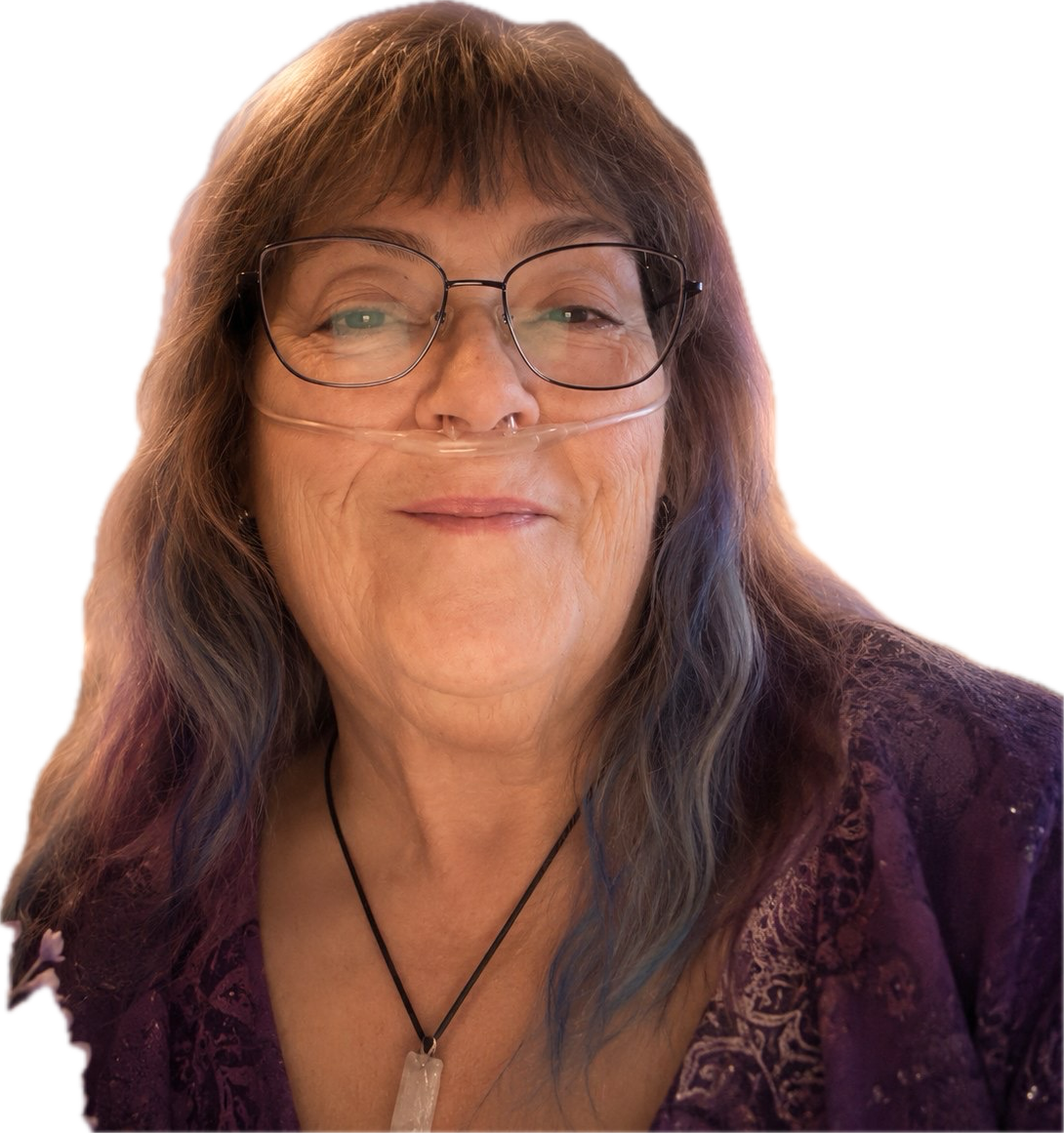 A woman with long, wavy hair and glasses, wearing a nasal cannula, smiling, dressed in a purple top, with a black necklace that has a white pendant.