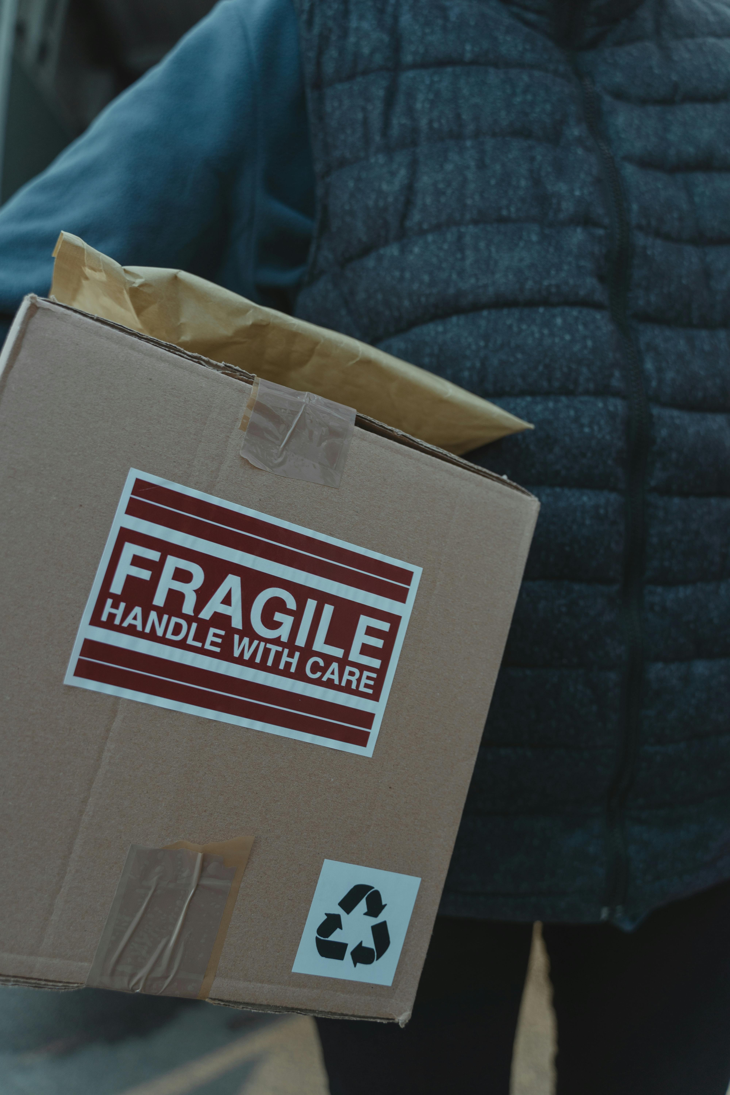 Person holding a cardboard box with a fragile sticker and a recycling symbol. The person is wearing a dark blue puffer jacket and dark pants.