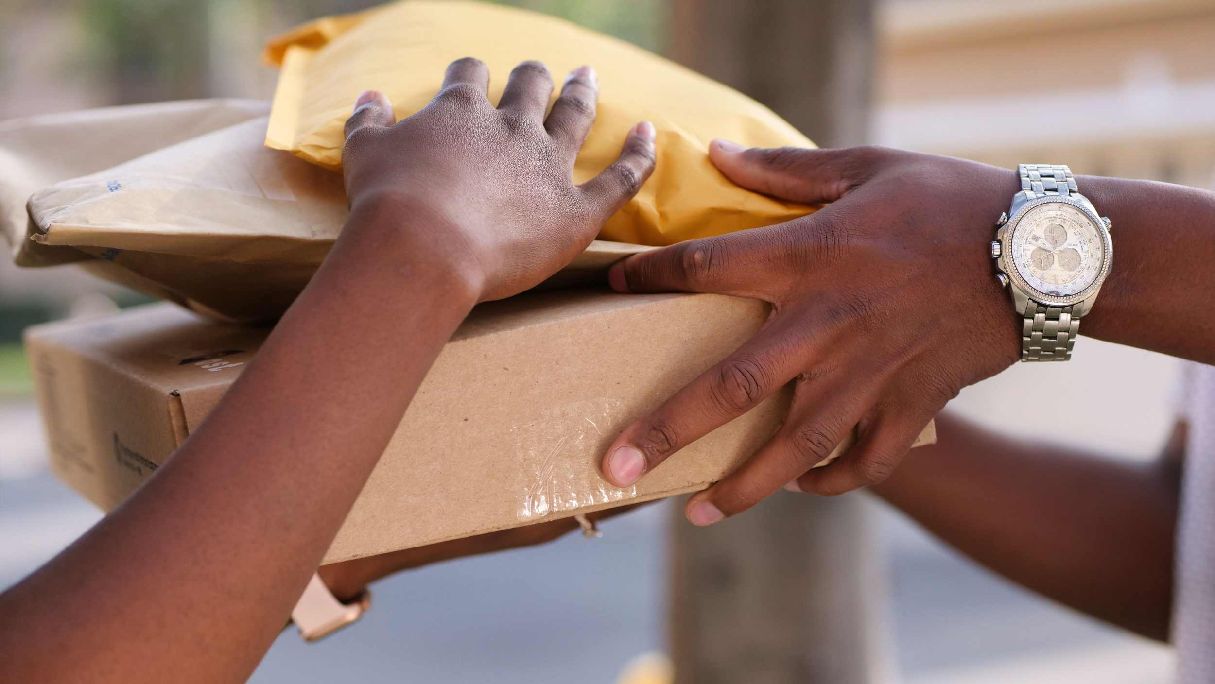Two people hands holding a cardboard box with a yellow padded envelope on top, with a blurred outdoor background.