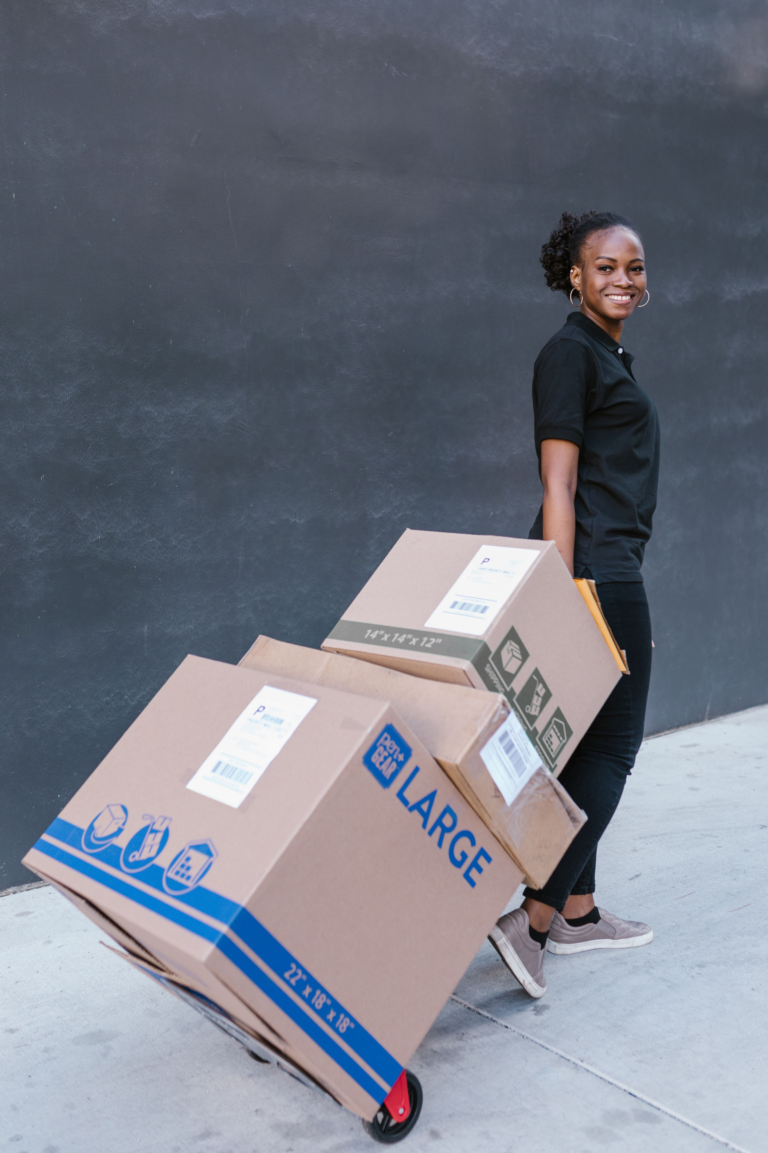 A woman smiling and standing next to a teal-colored hand truck carrying large cardboard boxes labeled for large office supplies, with black chalkboard background.