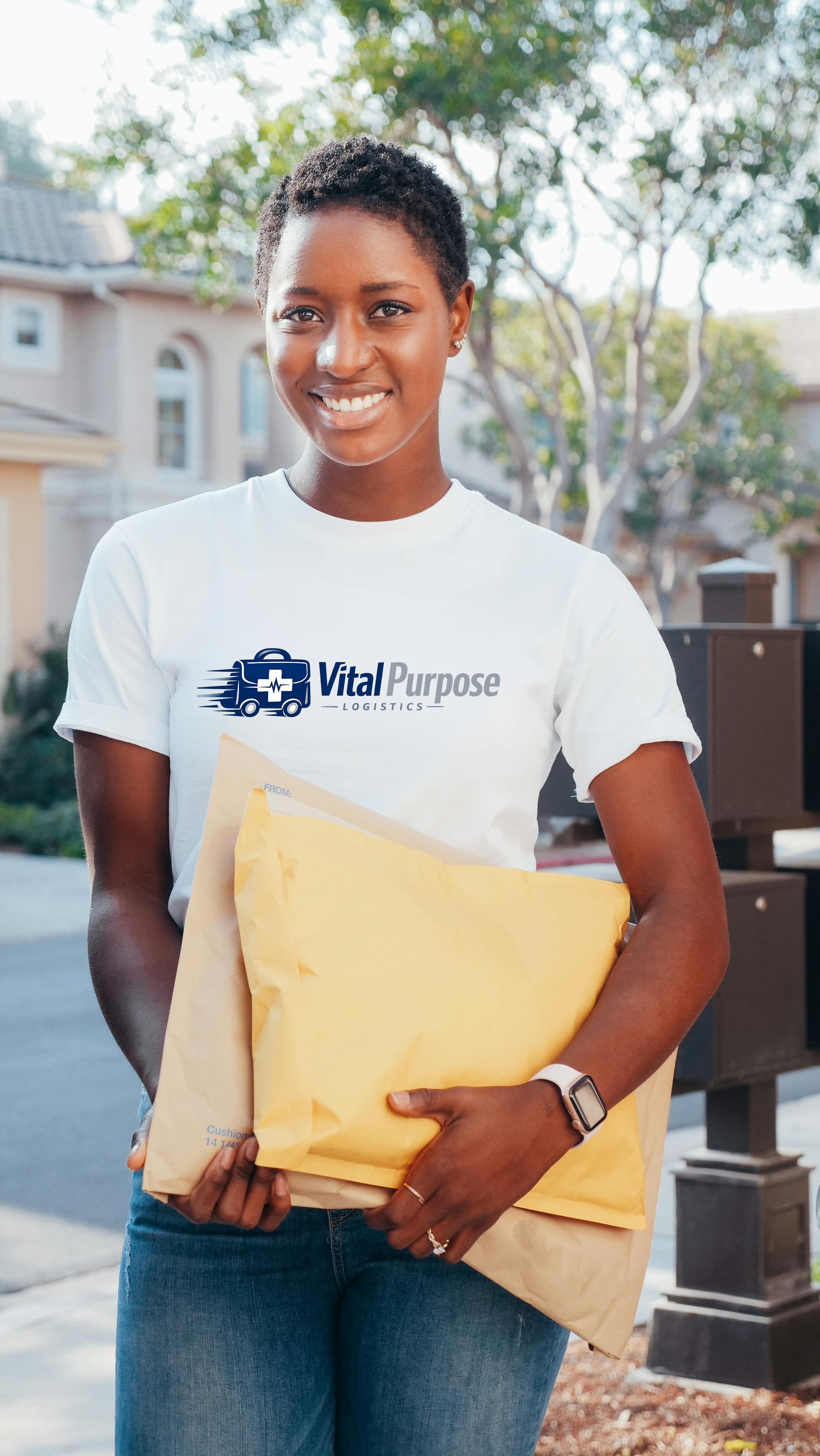 A smiling woman in a white T-shirt with Vital Purpose Logistics logo, holding a large yellow envelope, standing outdoors with trees and houses in the background.