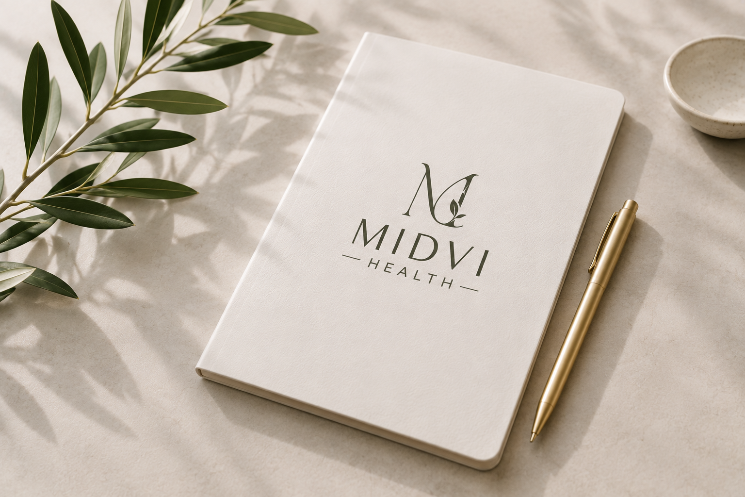 A white notebook with the logo and text 'MIDVI HEALTH' on the cover, a gold pen beside it, a small ceramic bowl, and a green leafy branch on a light-colored surface.