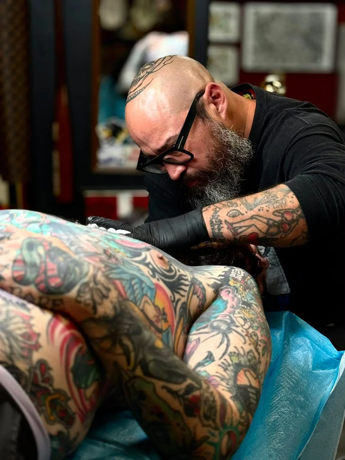 A man with tattoos, a beard, and short hair is standing indoors, wearing a sweatshirt that reads 'Maintain Your Business' with graffiti-style art, and looking to the right.