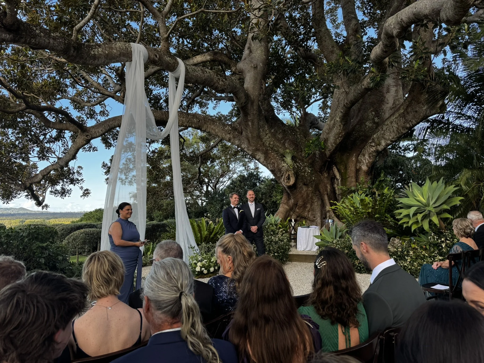 Outdoor wedding ceremony taking place under a large tree decorated with white fabric. Two men in suits are standing at the altar, and a woman in a blue dress is standing to the side. Guests are seated and standing in front, with lush plants and a scenic landscape in the background.