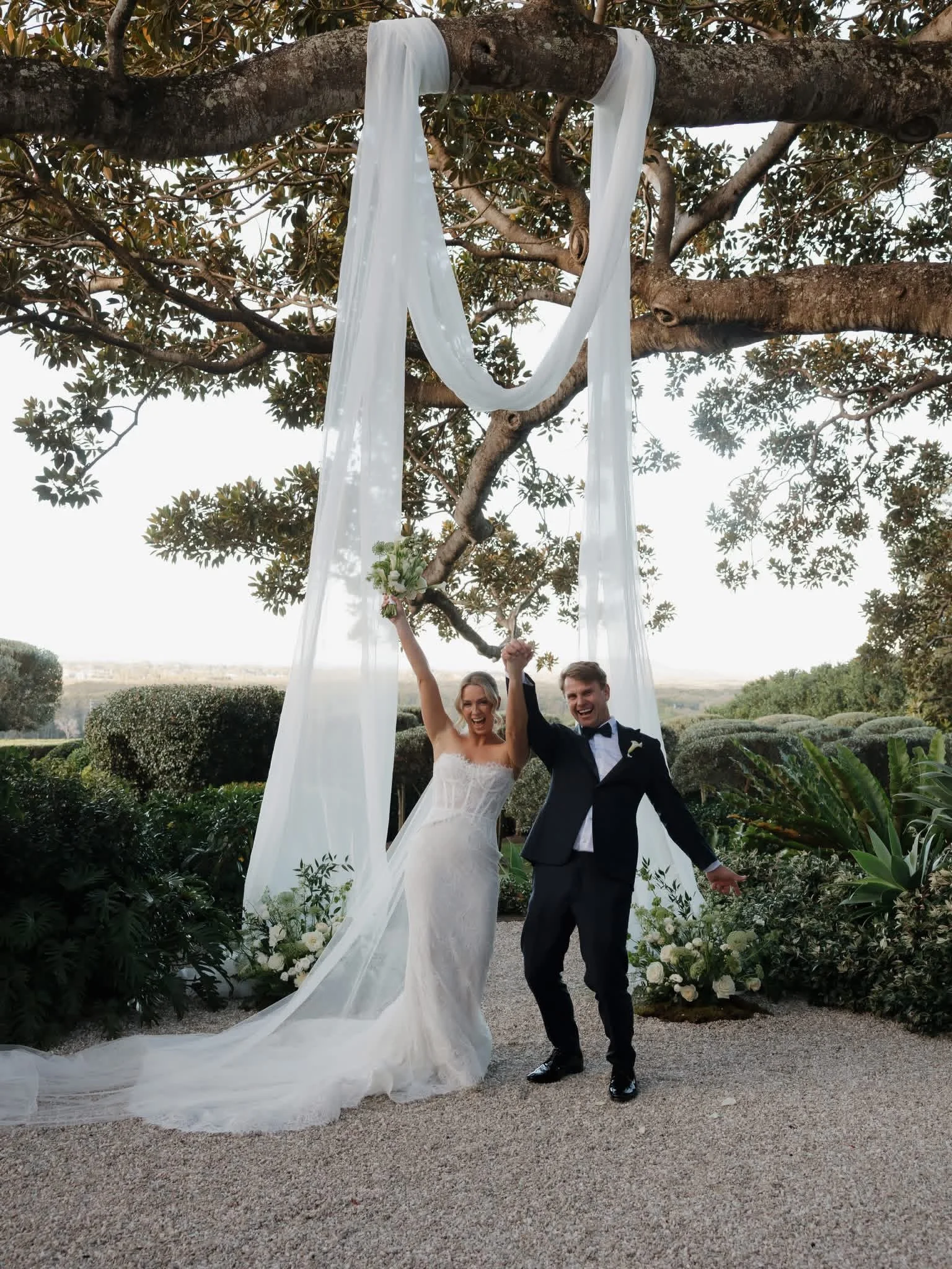 A newlywed couple dressed in wedding attire celebrating outdoors, with the bride in a white wedding dress and the groom in a tuxedo, standing beneath a large tree with white fabric draped from the branches, holding hands and smiling.