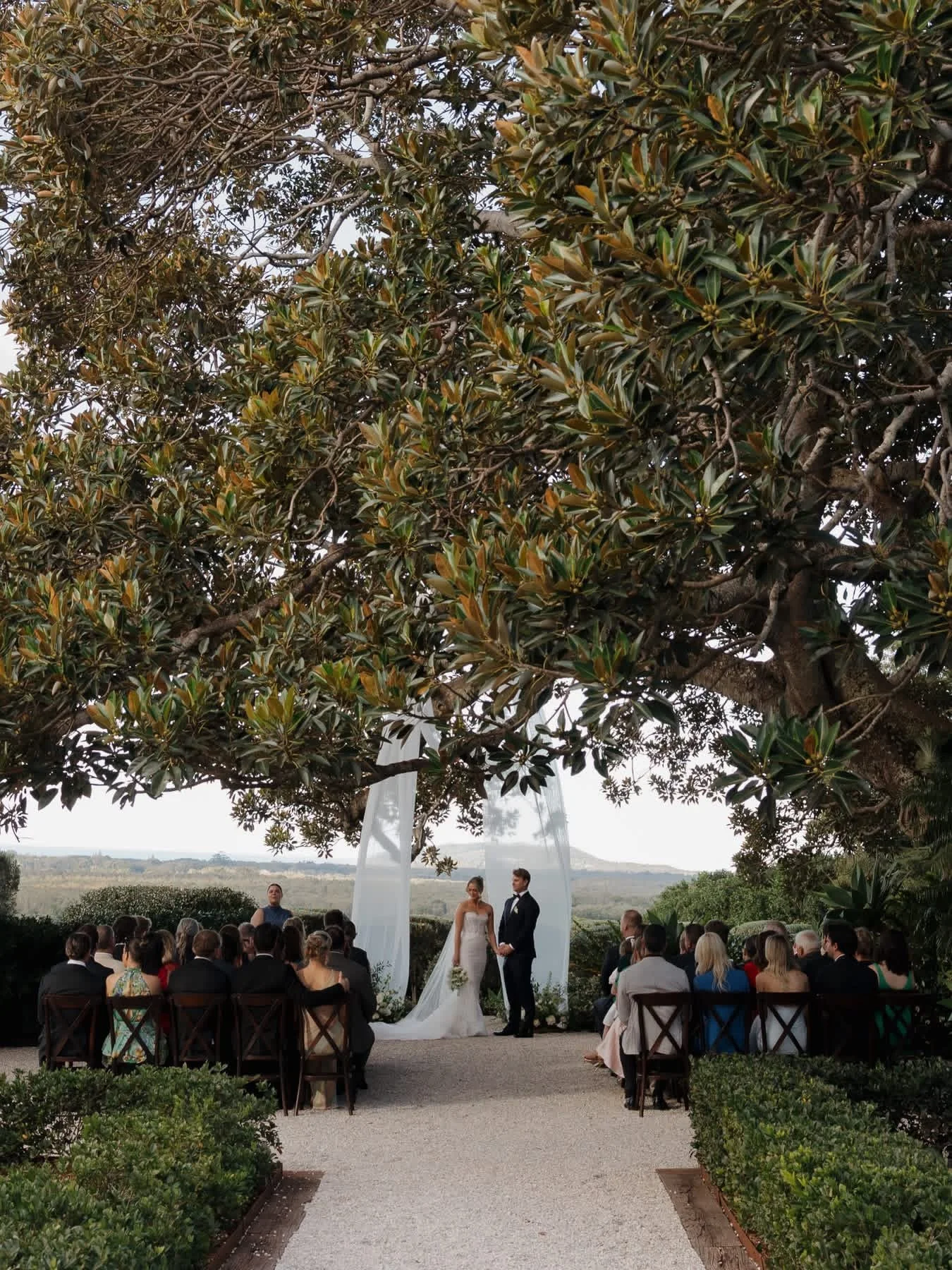 A couple getting married stands under a white fabric arch in an outdoor wedding ceremony, surrounded by guests seated in chairs, with a large leafy tree overhead and a landscape in the background.