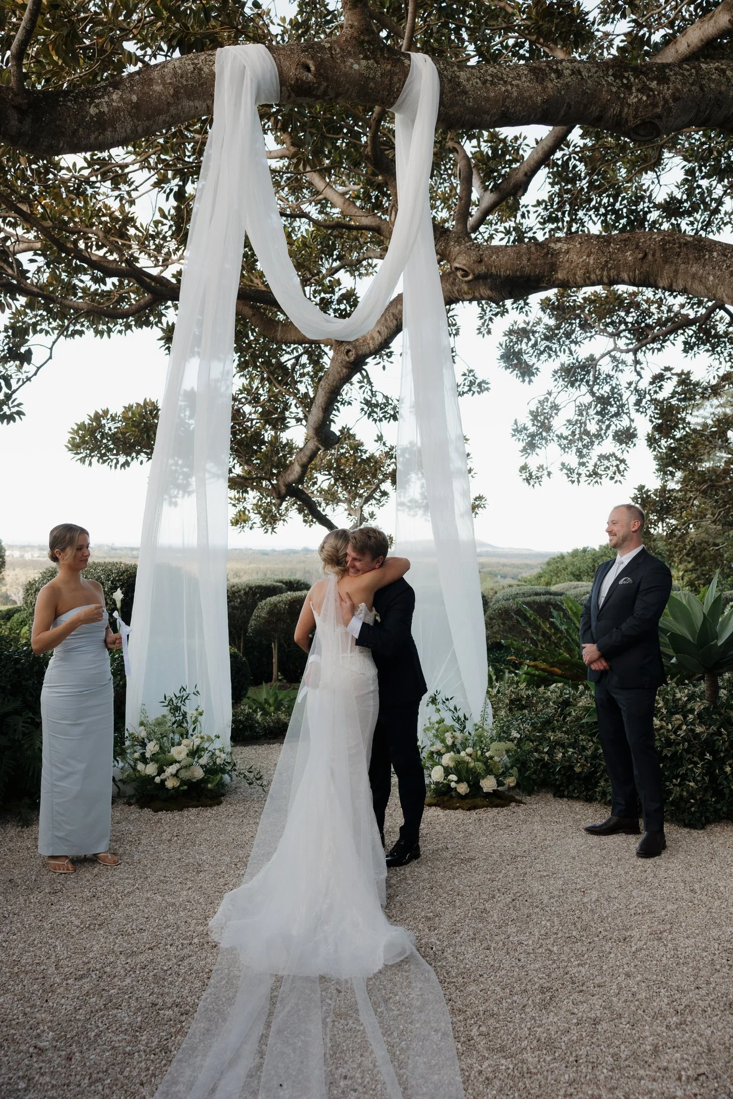 A couple is embracing under a large tree decorated for a wedding, with white curtains hanging from the branches. There are two women and a man standing nearby, all dressed in formal attire, in a garden setting with greenery and flowers.