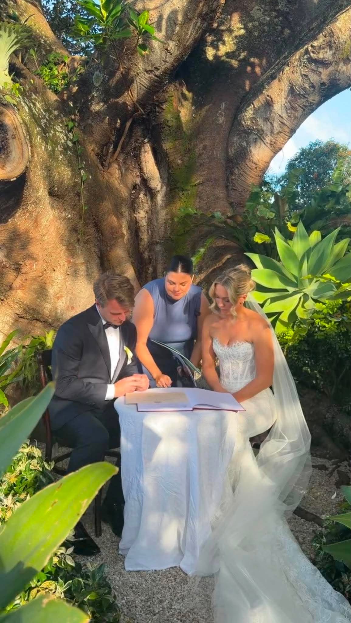A bride and groom sitting at a table outdoors under a large tree, signing a document during a wedding ceremony, with a woman helping them.