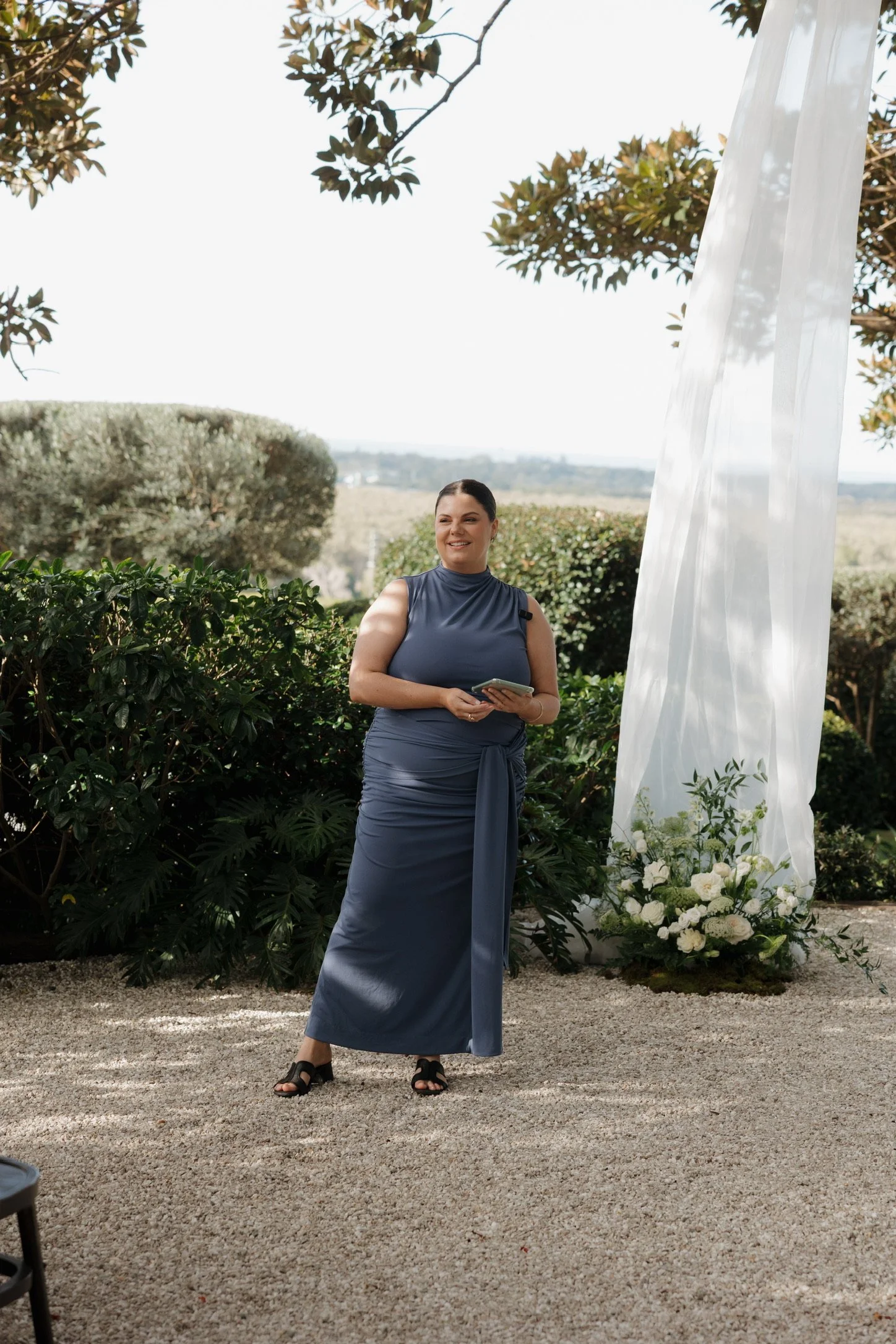 A woman in a blue dress and black sandals standing outdoors surrounded by bushes, white flowers, and trees, with a white fabric backdrop and a scenic background of open land and hills.