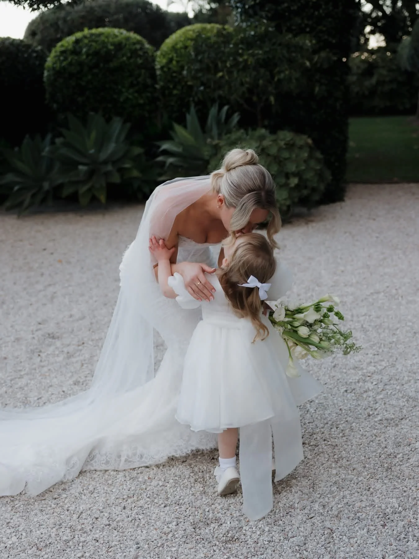 A bride and a young girl, possibly a flower girl, share a joyful moment outdoors. The bride is leaning down and kissing the girl, who is holding a bouquet of white flowers. The bride is wearing a white wedding gown with a veil, and the girl is dressed in a white dress with a ribbon in her hair. The background features lush green bushes and trees.