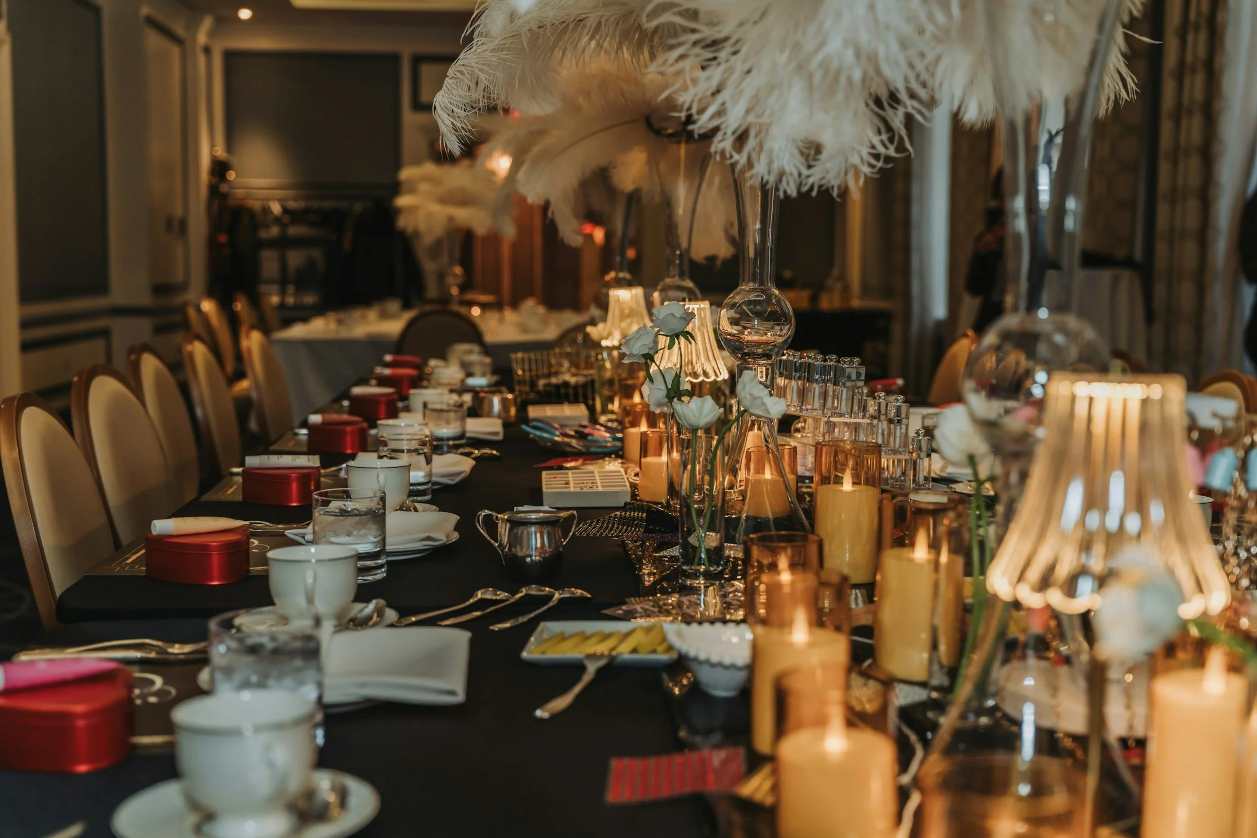 Elegant banquet table decorated with white flowers, tall feather centerpieces in glass vases, and various tableware in a dimly lit room prepared for a celebration or event.