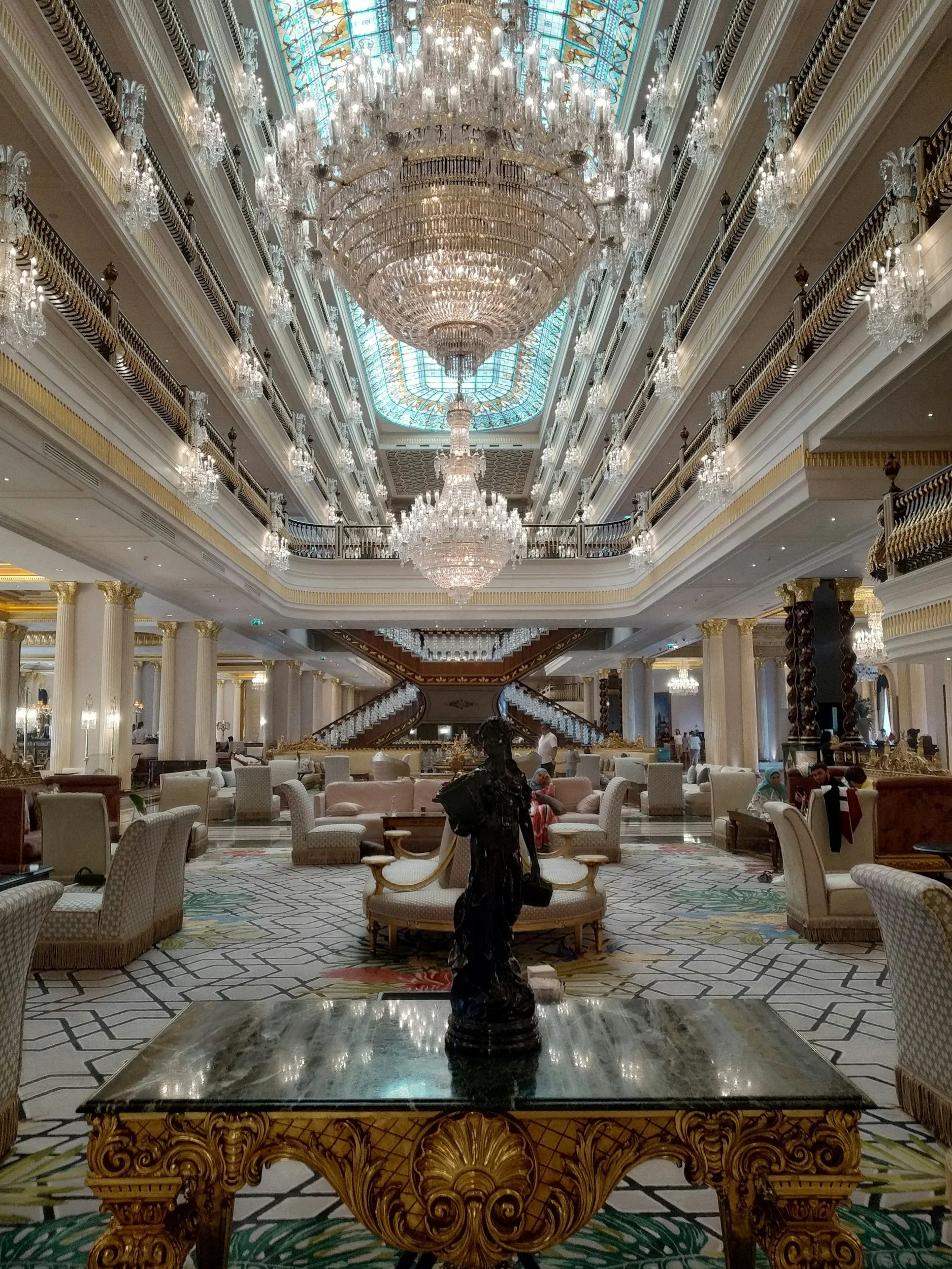 Elegant hotel lobby with large crystal chandeliers, ornate columns, luxurious seating, and a stained glass ceiling. There are people sitting and walking around.