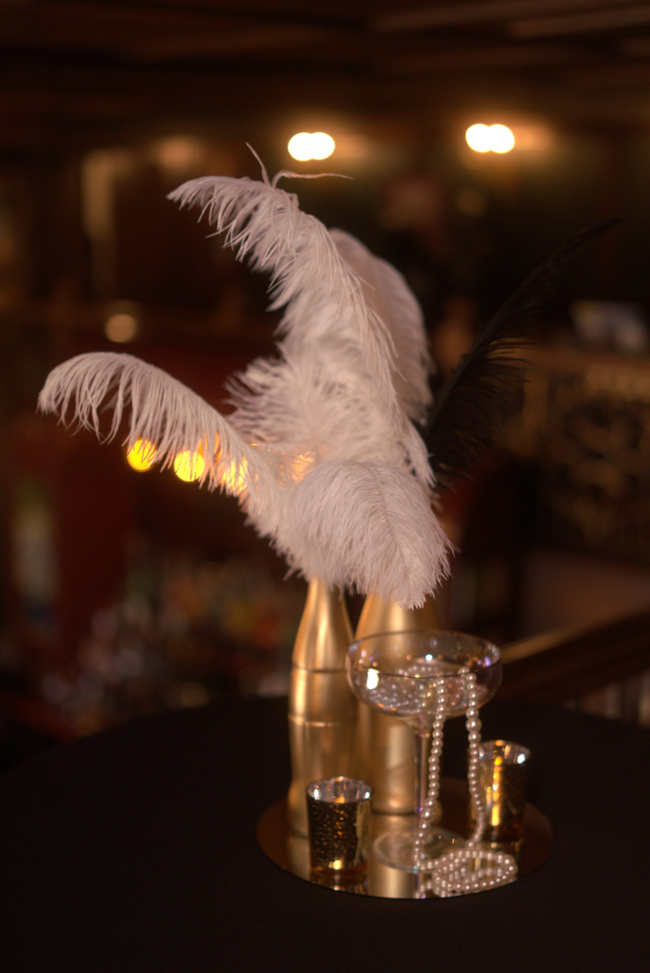 Close-up of a feathered decoration on a table with gold cups and jewelry, blurred lights in the background.