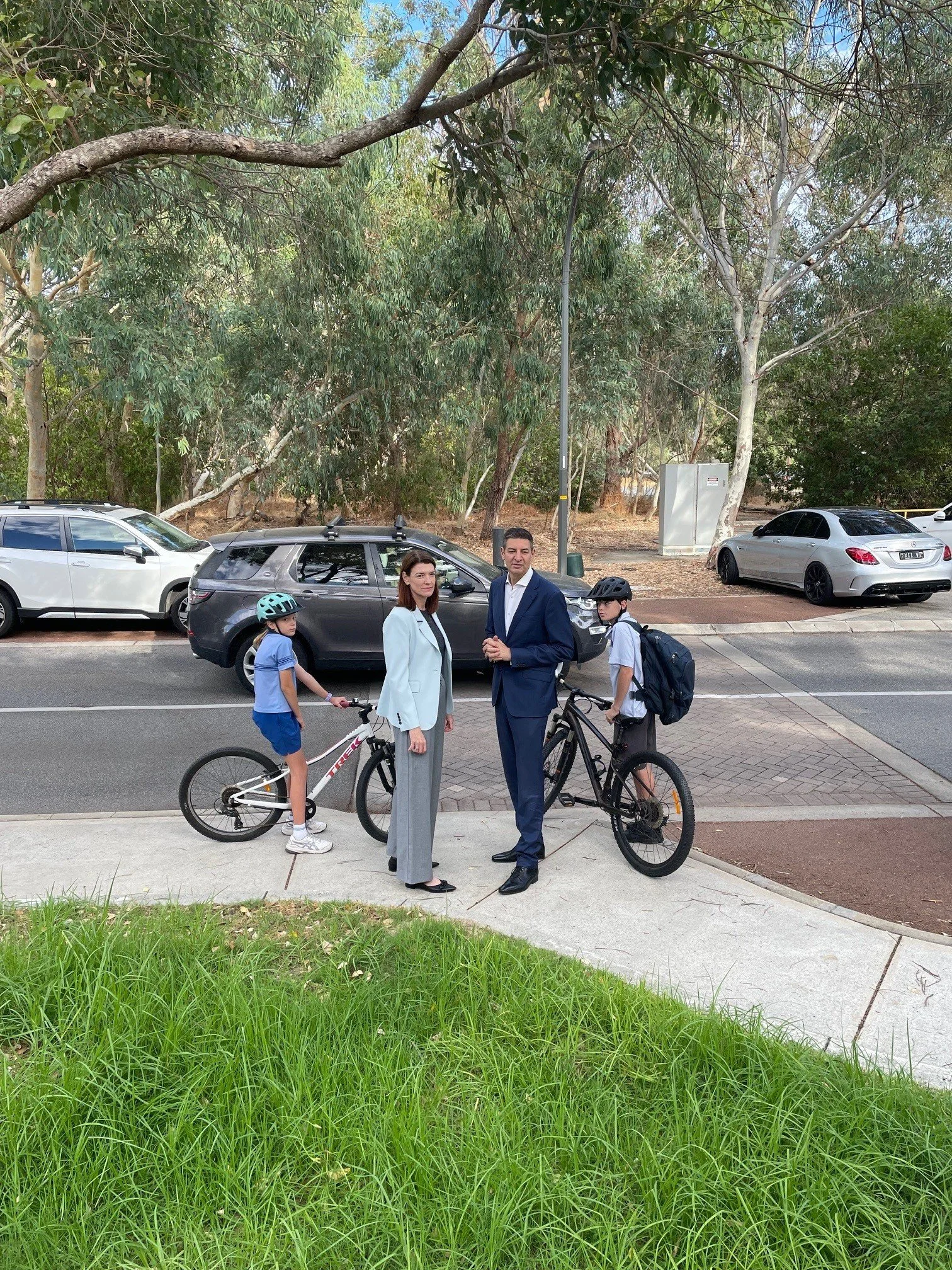 A man, a woman, and two children with bikes standing on a sidewalk in front of a parking lot with parked cars, trees, and greenery.