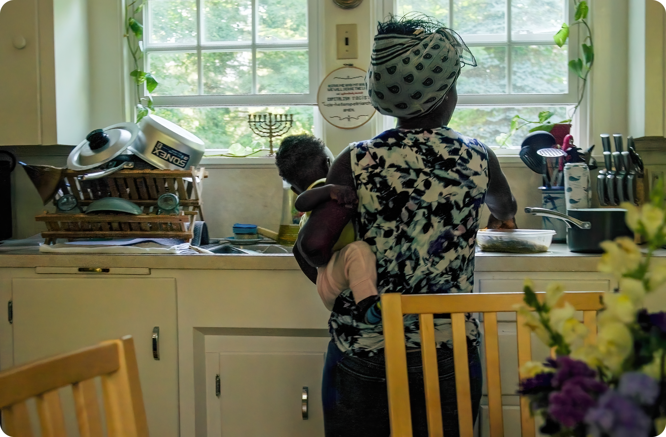 A woman wearing a patterned headscarf and floral shirt holds a child in a kitchen, facing a window with green trees outside.