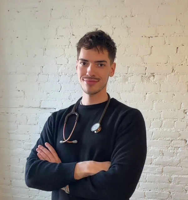 Young male healthcare professional standing with arms crossed, wearing a stethoscope around his neck in front of a white brick wall.