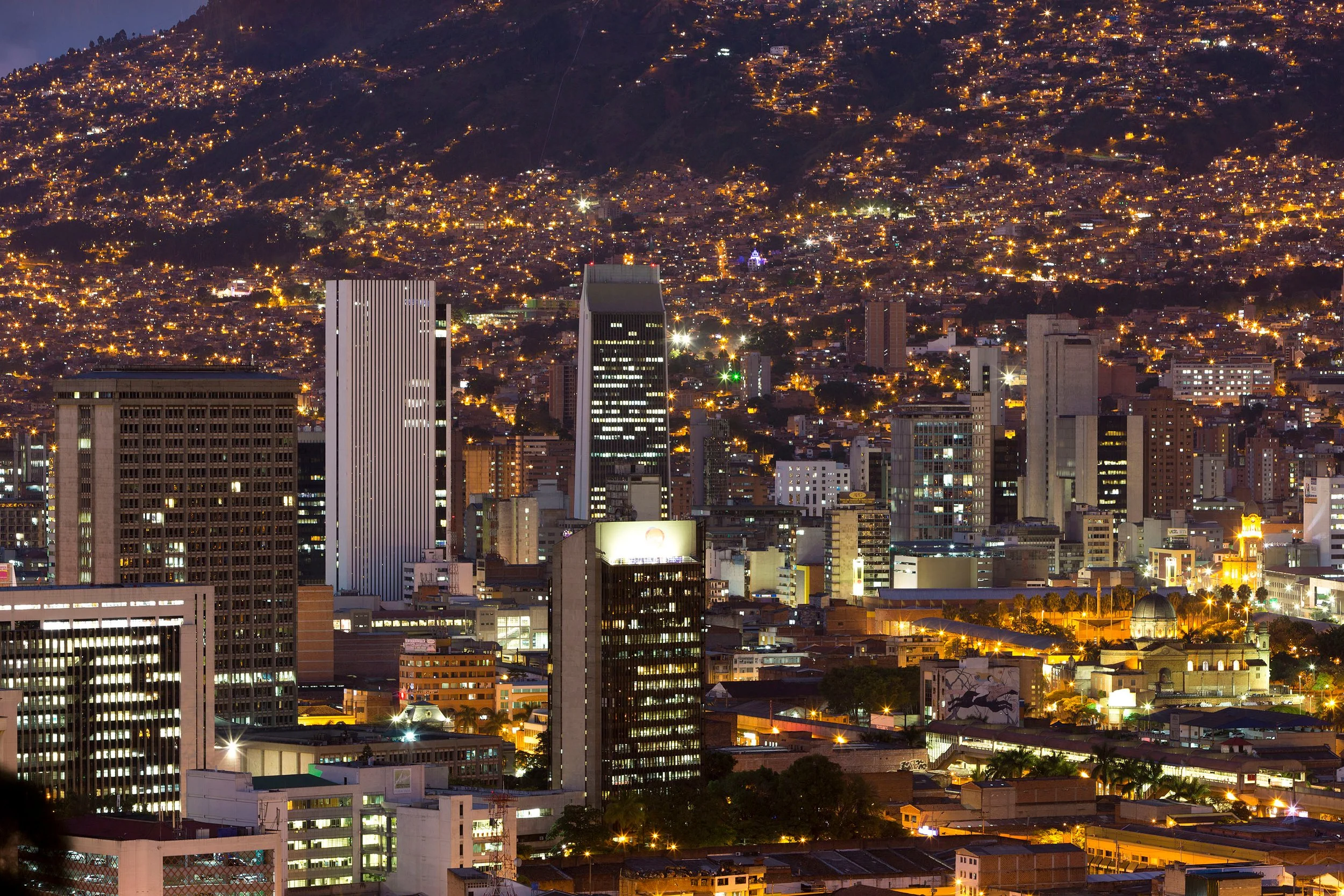 Vista nocturna de una ciudad con numerosos edificios y luces, con colinas en el fondo y un cielo oscuro.