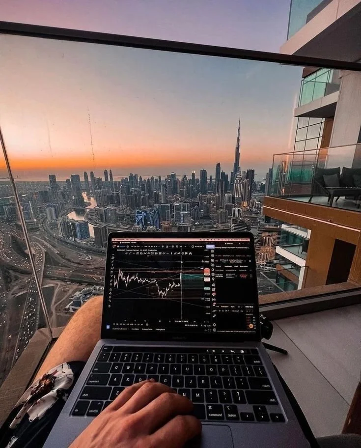 A person working on a laptop with a city skyline view of Dubai including the Burj Khalifa at sunset from a high-rise apartment.