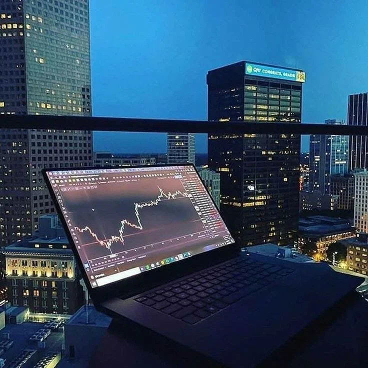 A laptop on a balcony overlooking a cityscape at dusk or night, with a stock chart displayed on the screen and tall office buildings in the background.