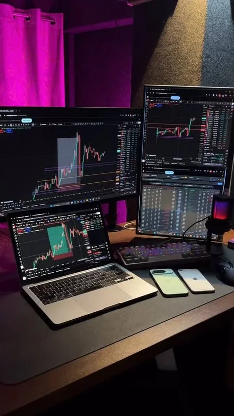 Multiple computer screens displaying stock or cryptocurrency trading charts in a dimly lit room with purple curtains.