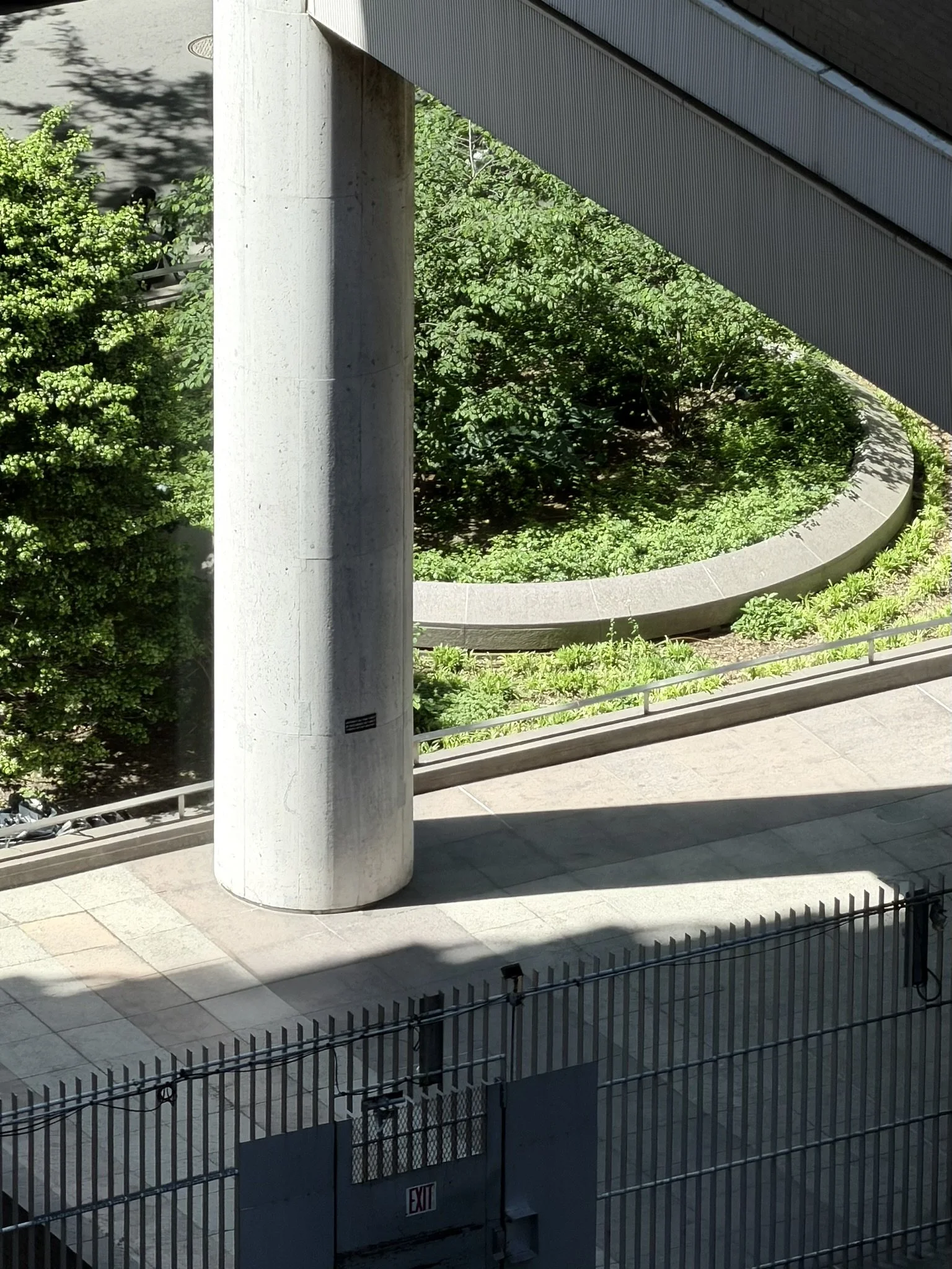 A view of a parking area and a landscaped garden with green bushes and trees, separated by a concrete walkway and fencing.
