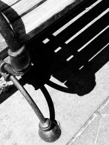Close-up of a wooden park bench with cast iron armrest and shadow on concrete sidewalk in black and white.