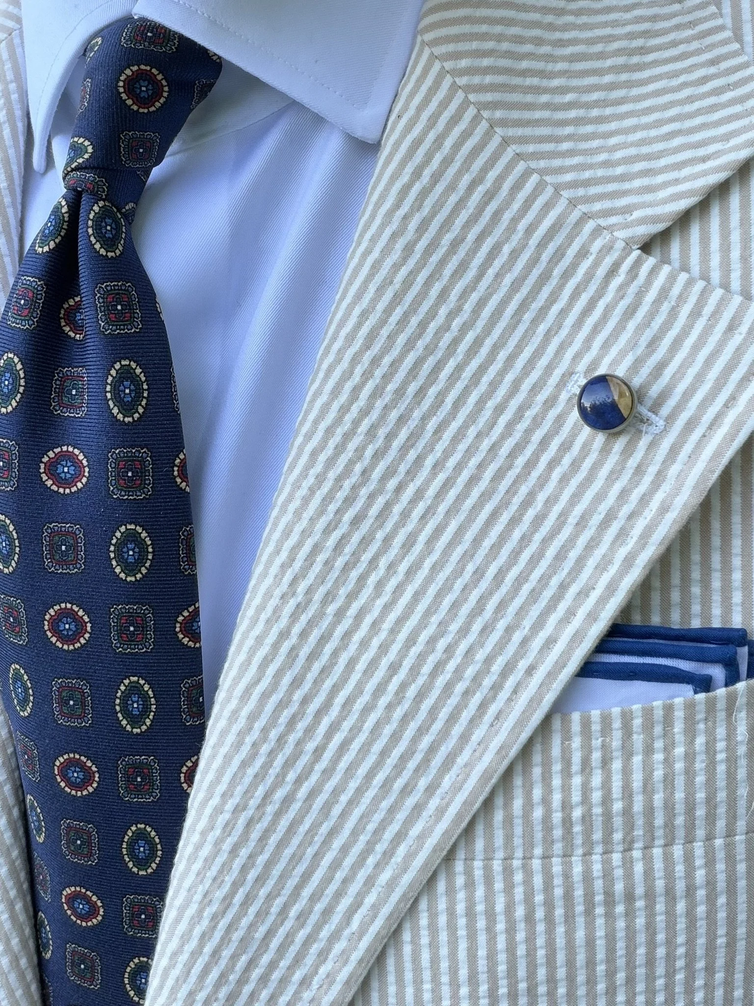Close-up of a man wearing a cream and white striped blazer, a white shirt, a patterned blue tie, a blue and yellow striped pocket square, and a round lapel pin.
