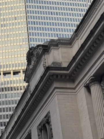 Close-up of a historical building with classical architectural features, including columns and decorative details, against a backdrop of modern high-rise office buildings.