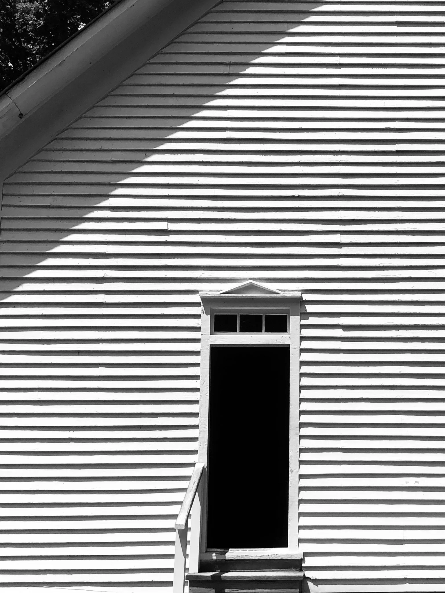 Black and white photo of an empty house entrance with staircase leading up to an un-paneled door. The house has horizontal wooden siding, and the scene is partially shadowed.